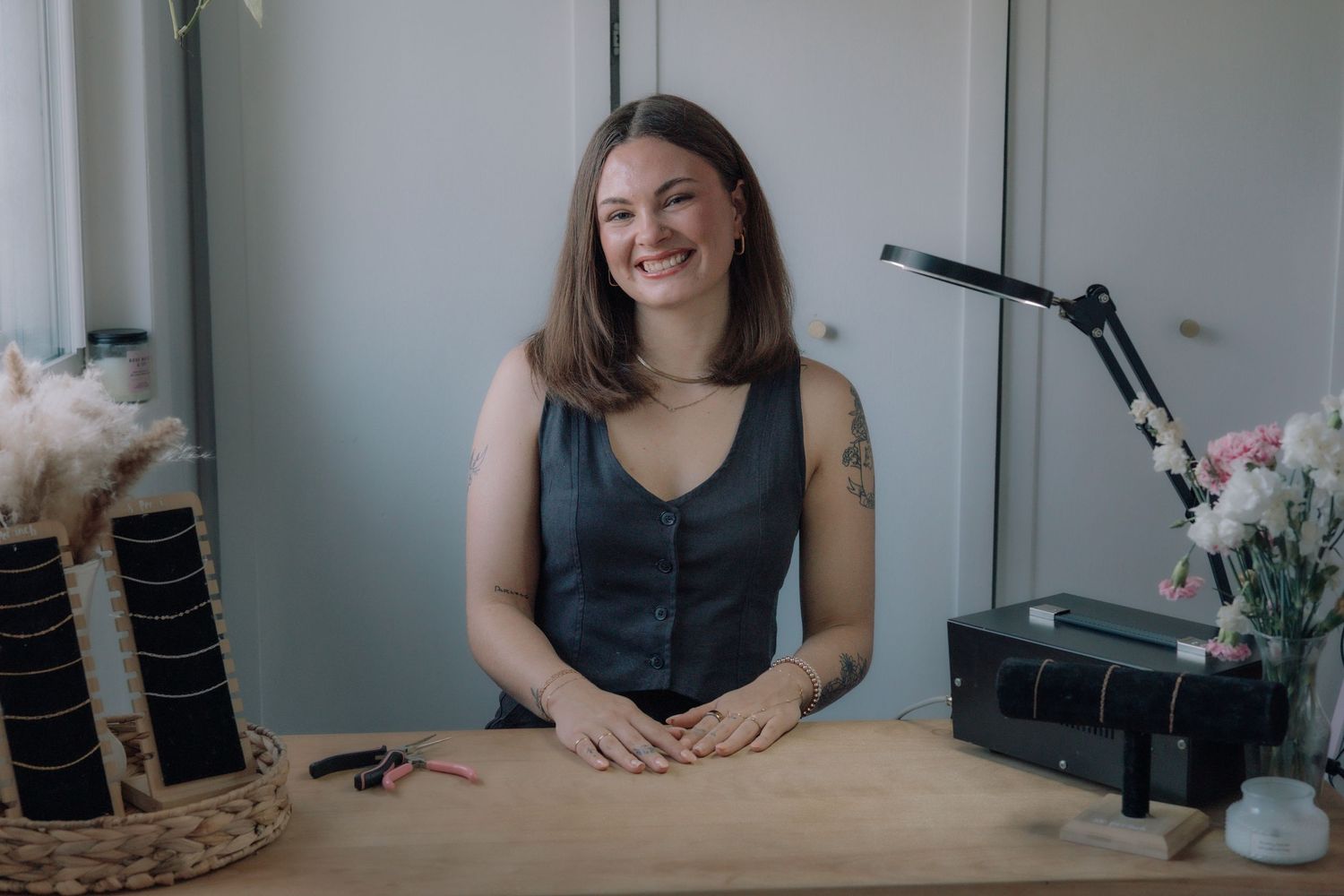 Smiling brunette woman sitting at a wooden desk in a bright studio with flowers in background.