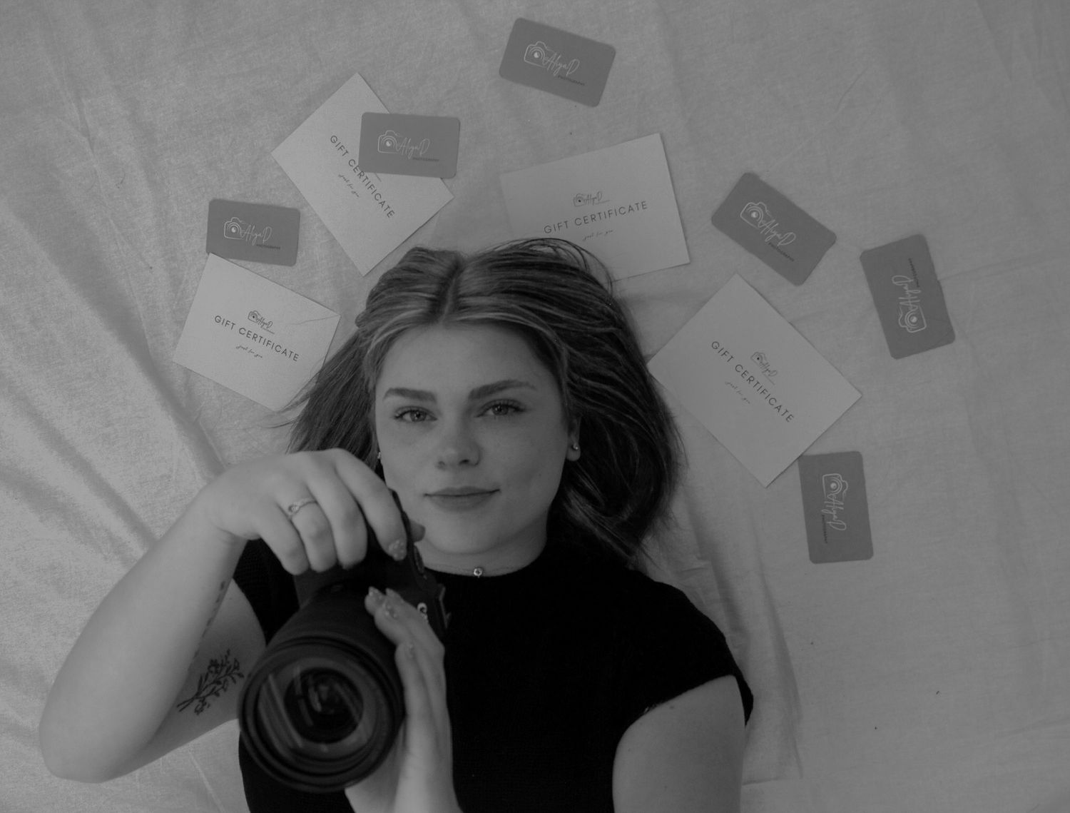 Black and white portrait of young woman lying down holding camera surrounded by cards.