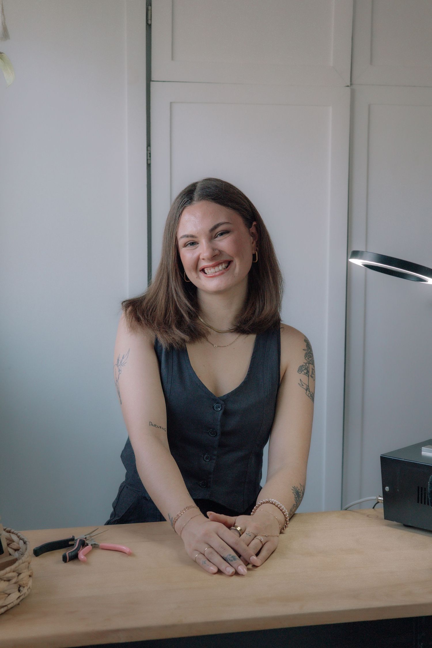 Smiling brunette woman leaning on a wooden desk in a gray sleeveless top with arm tattoos.