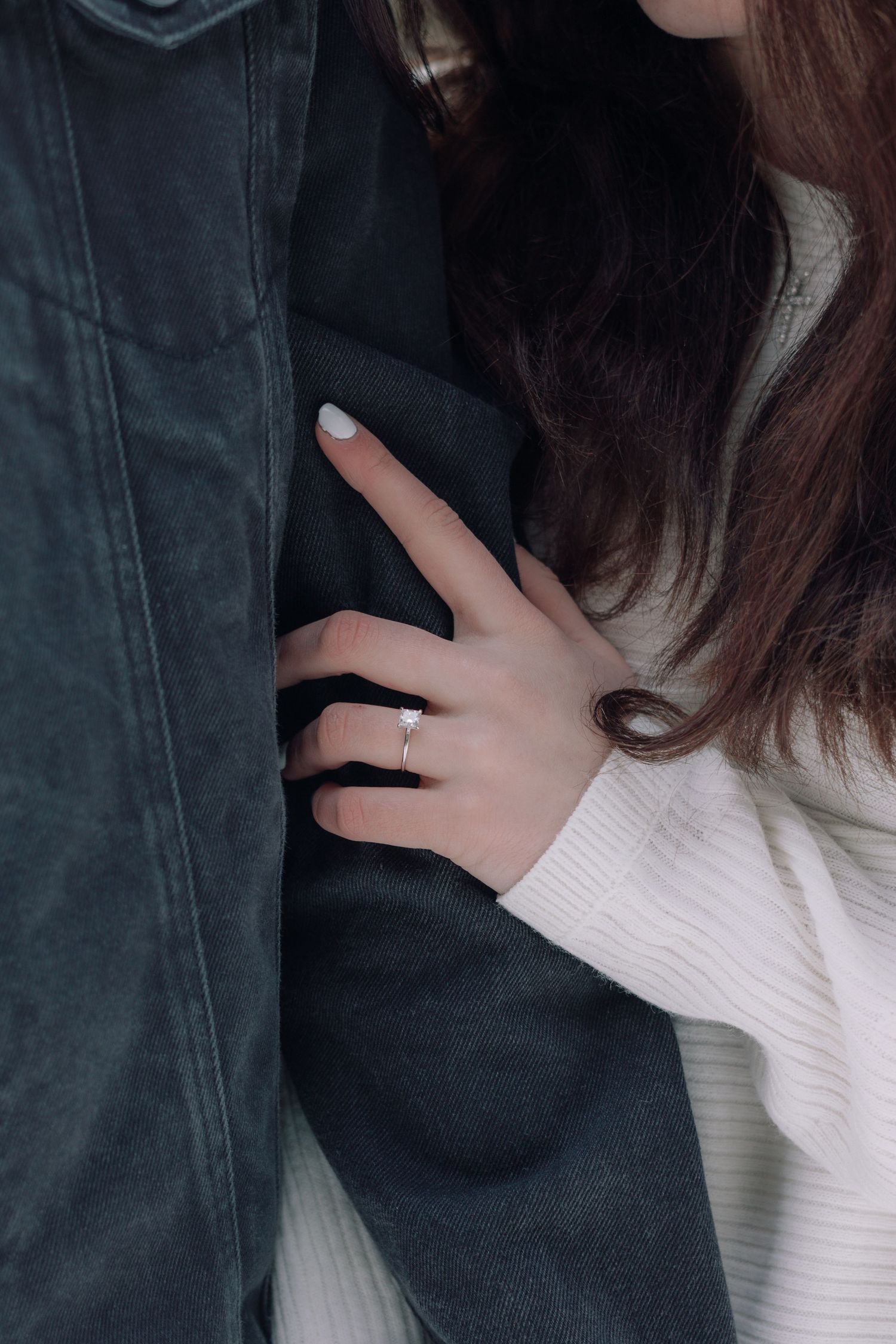 A close-up of a womans hand wearing a diamond engagement ring resting on a dark jacket.