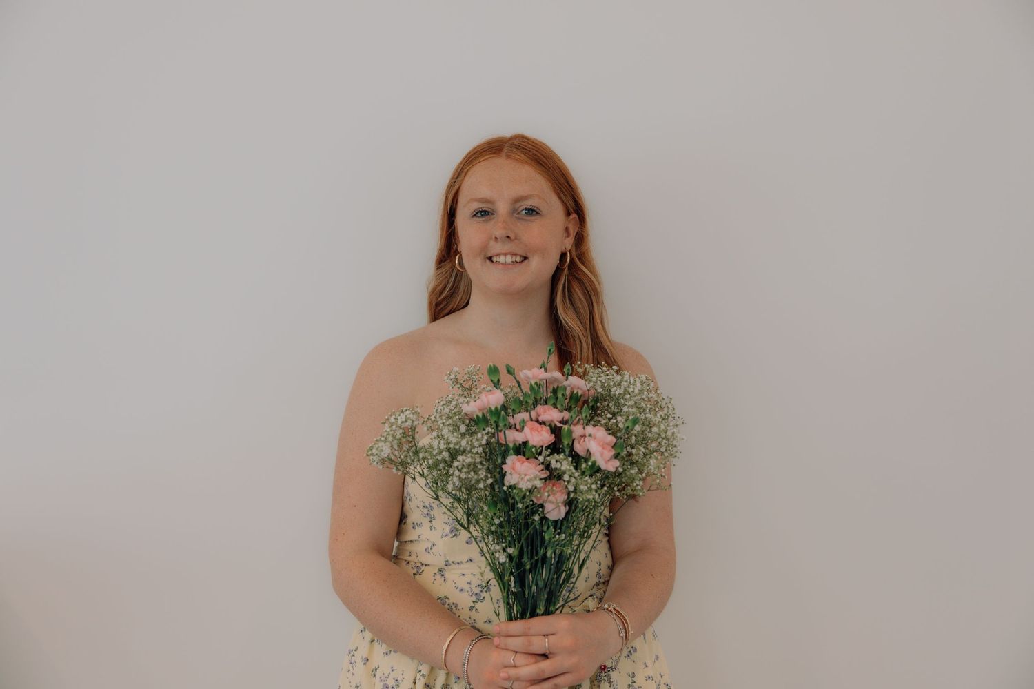 Smiling redhead woman holding a bouquet of pink carnations and baby's breath against a white wall.