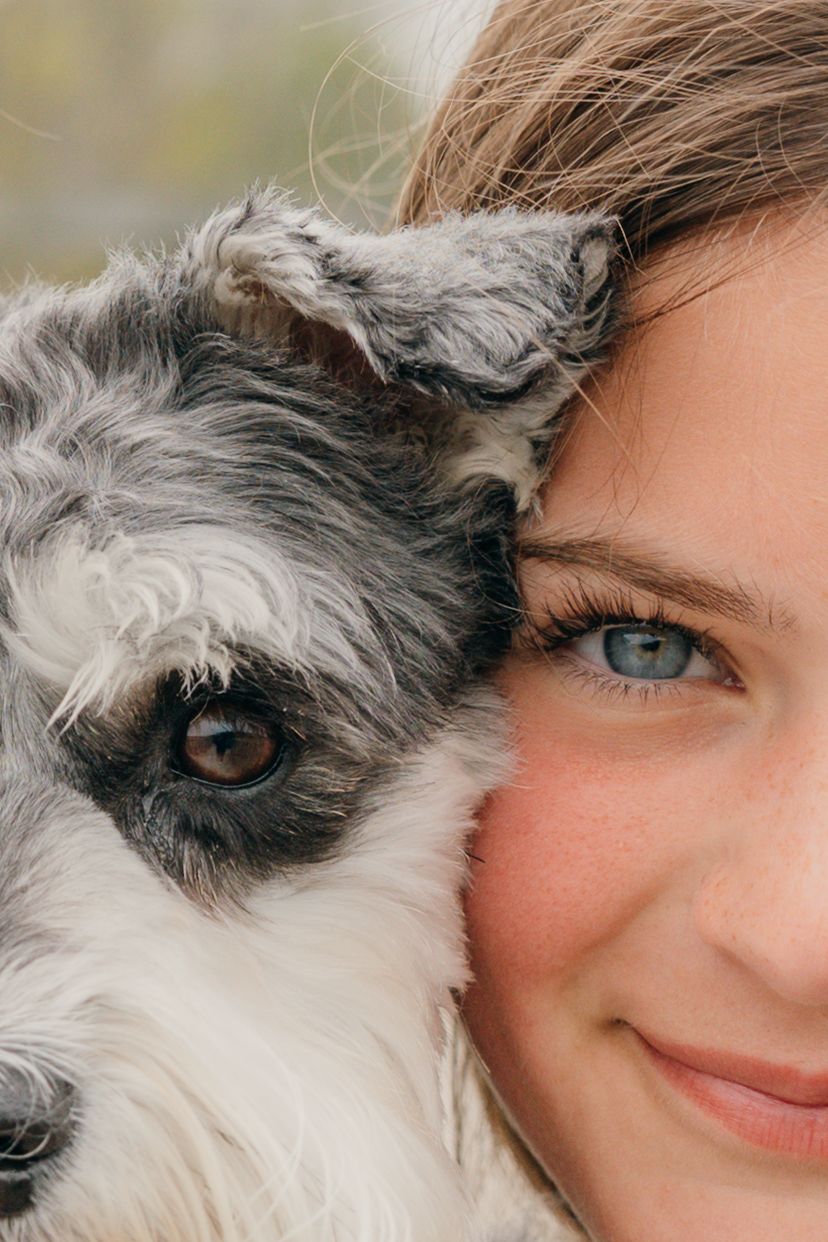 Close-up portrait of a smiling young woman cheek-to-cheek with a gray and white fluffy dog.
