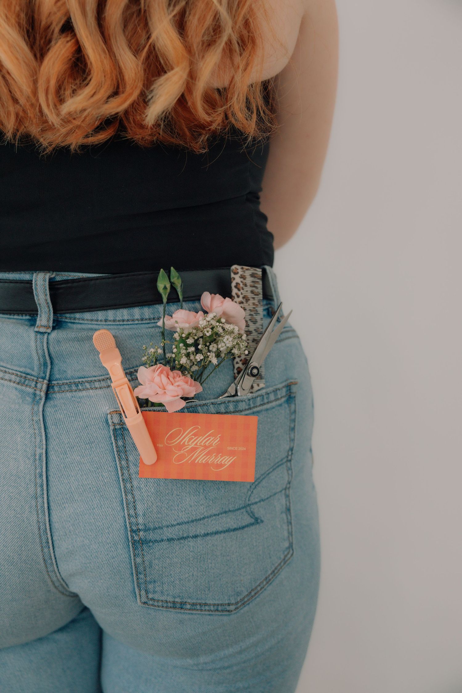 Back view of woman in jeans with hair styling tools, flowers, and business card in back pocket.