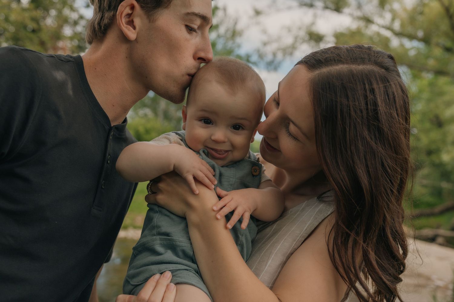 Two parents lovingly kiss their smiling baby on the cheeks outdoors in soft natural light.
