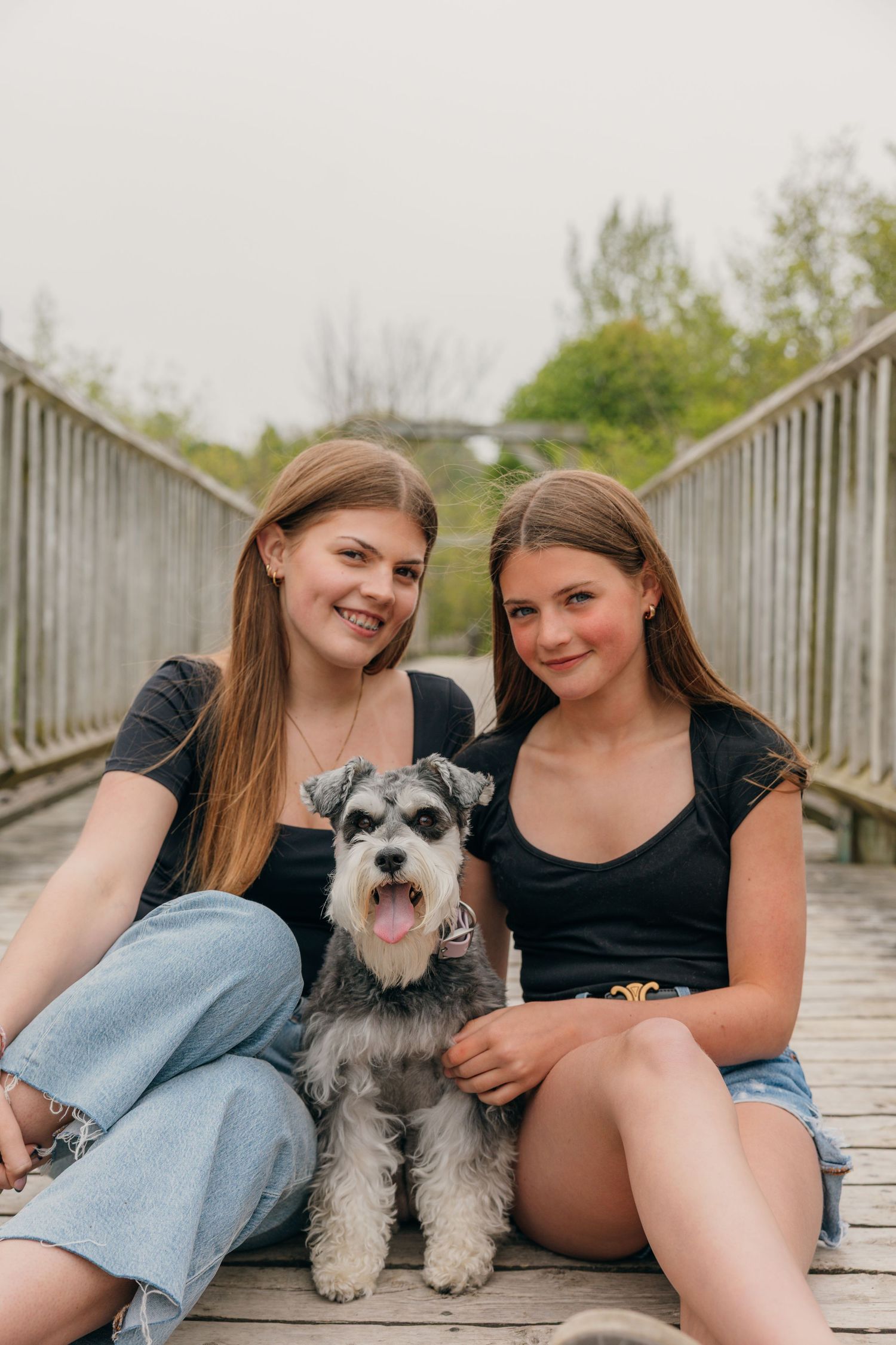 Two teenage girls sitting on wooden bridge boardwalk with miniature schnauzer dog.