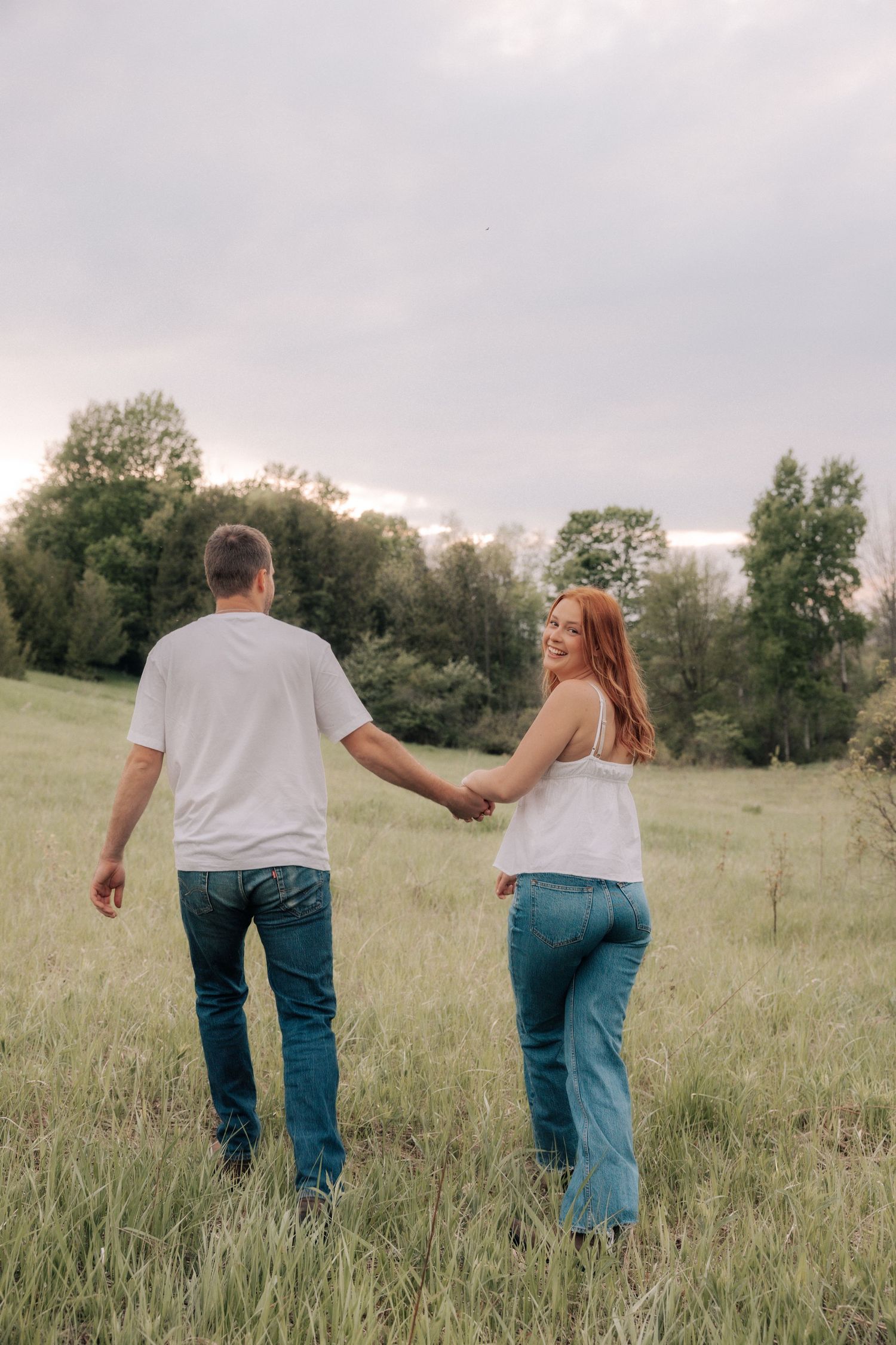 Happy couple holding hands walking through a green meadow under a cloudy sky.