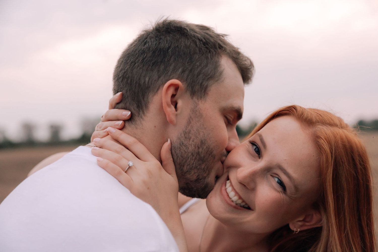 Smiling redhead woman embracing man kissing her cheek during romantic outdoor engagement session.