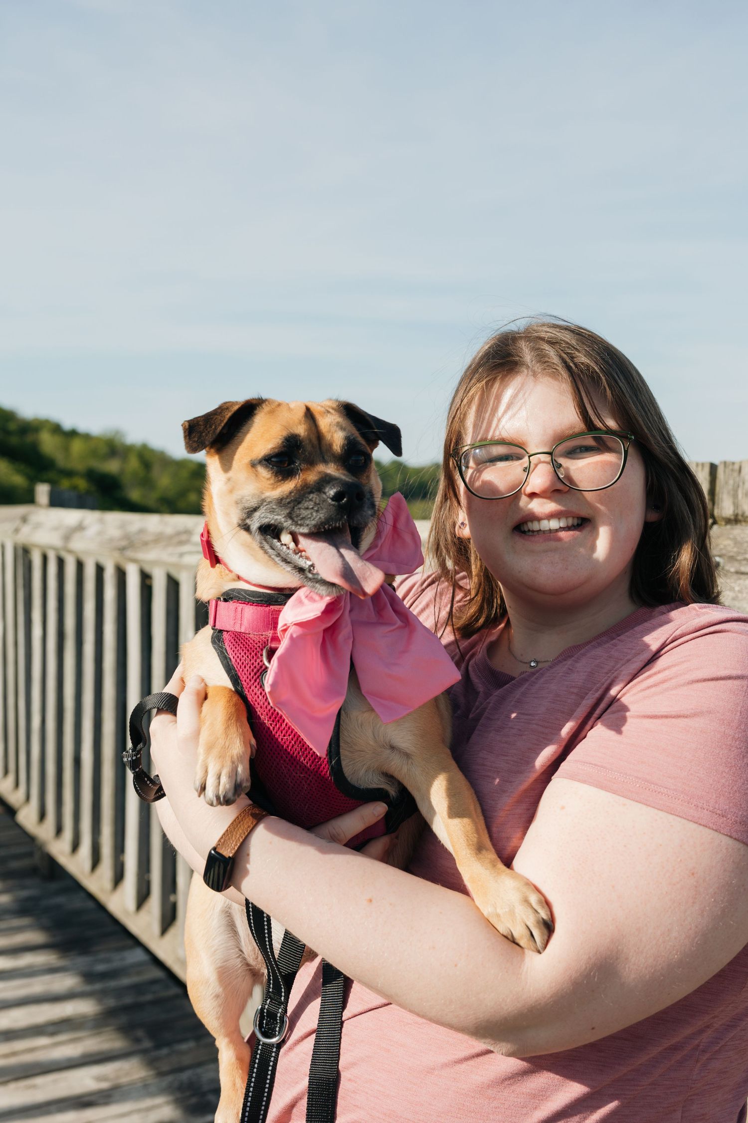 Smiling woman with glasses holding small brown dog with pink bow on sunny bridge.