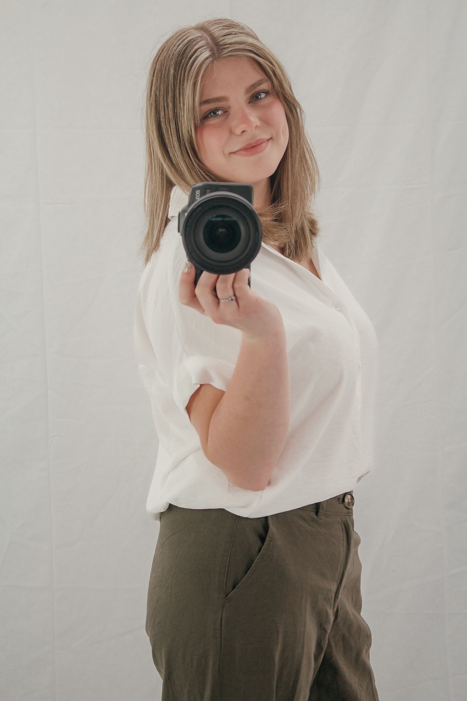 Young female photographer smiling and holding Canon DSLR camera against white fabric backdrop.