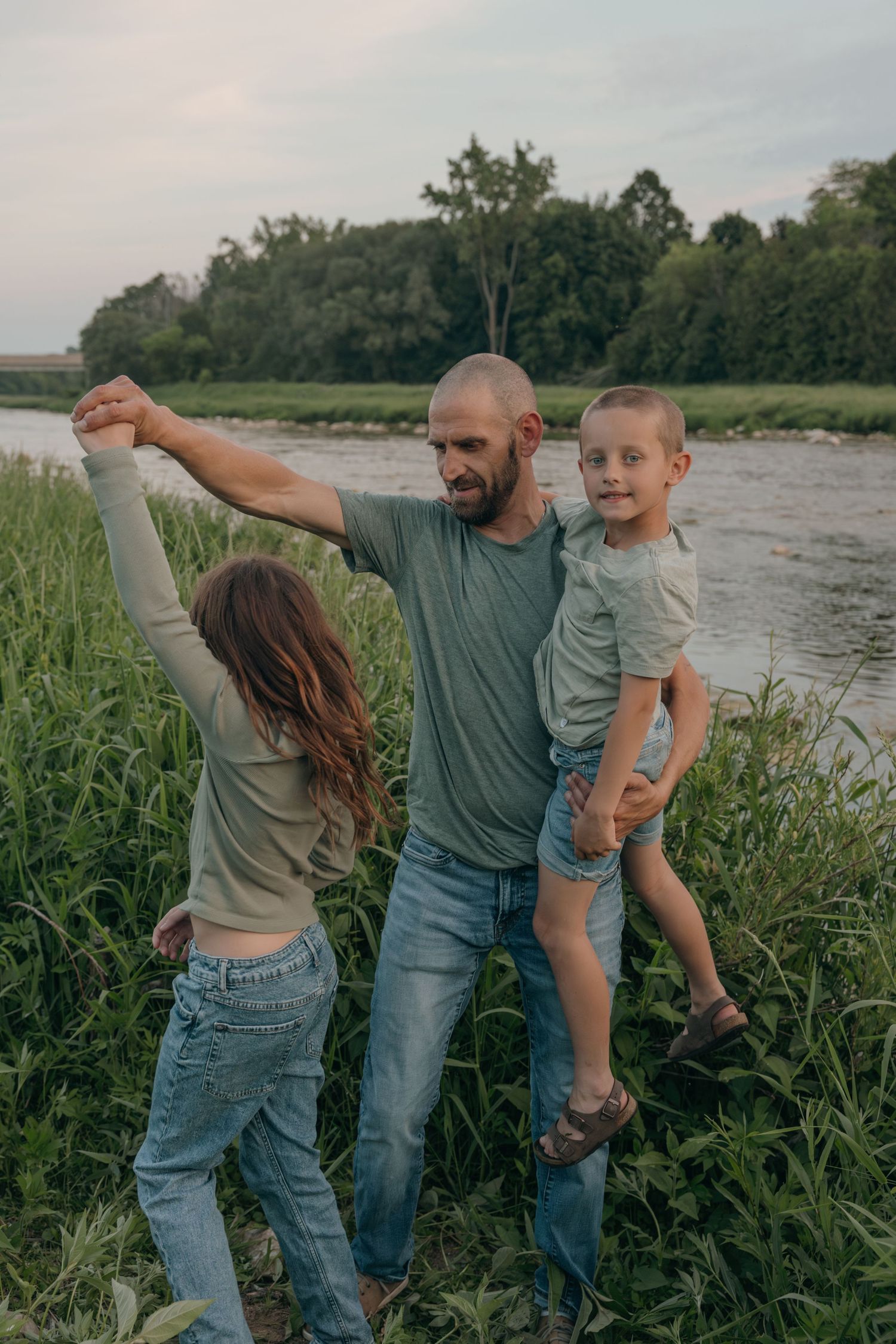 Father playfully holding son and spinning daughter by hand in a green riverside meadow at dusk.