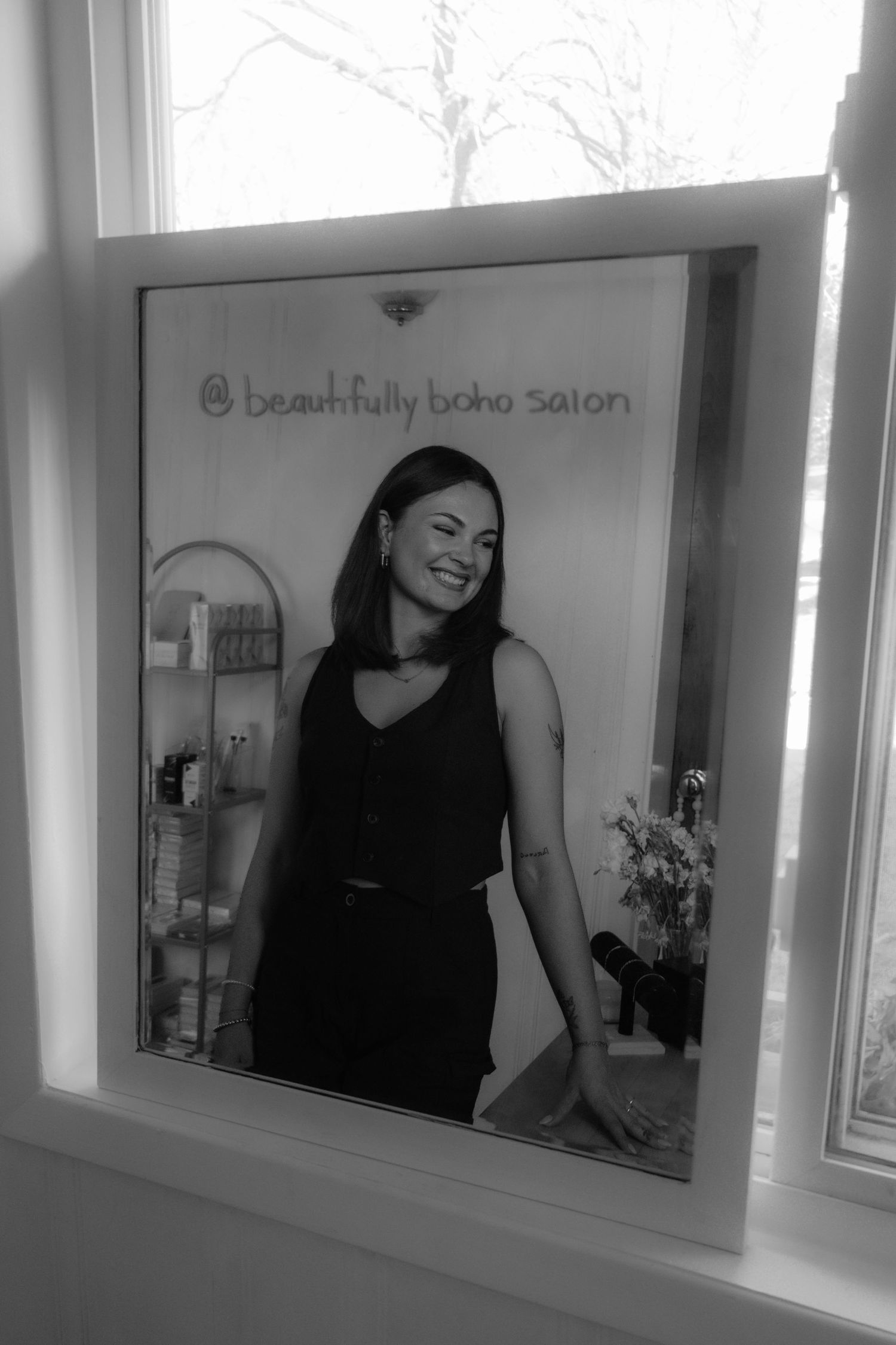 Black and white photo of a smiling woman in a window display for Beautifully Boho Salon.