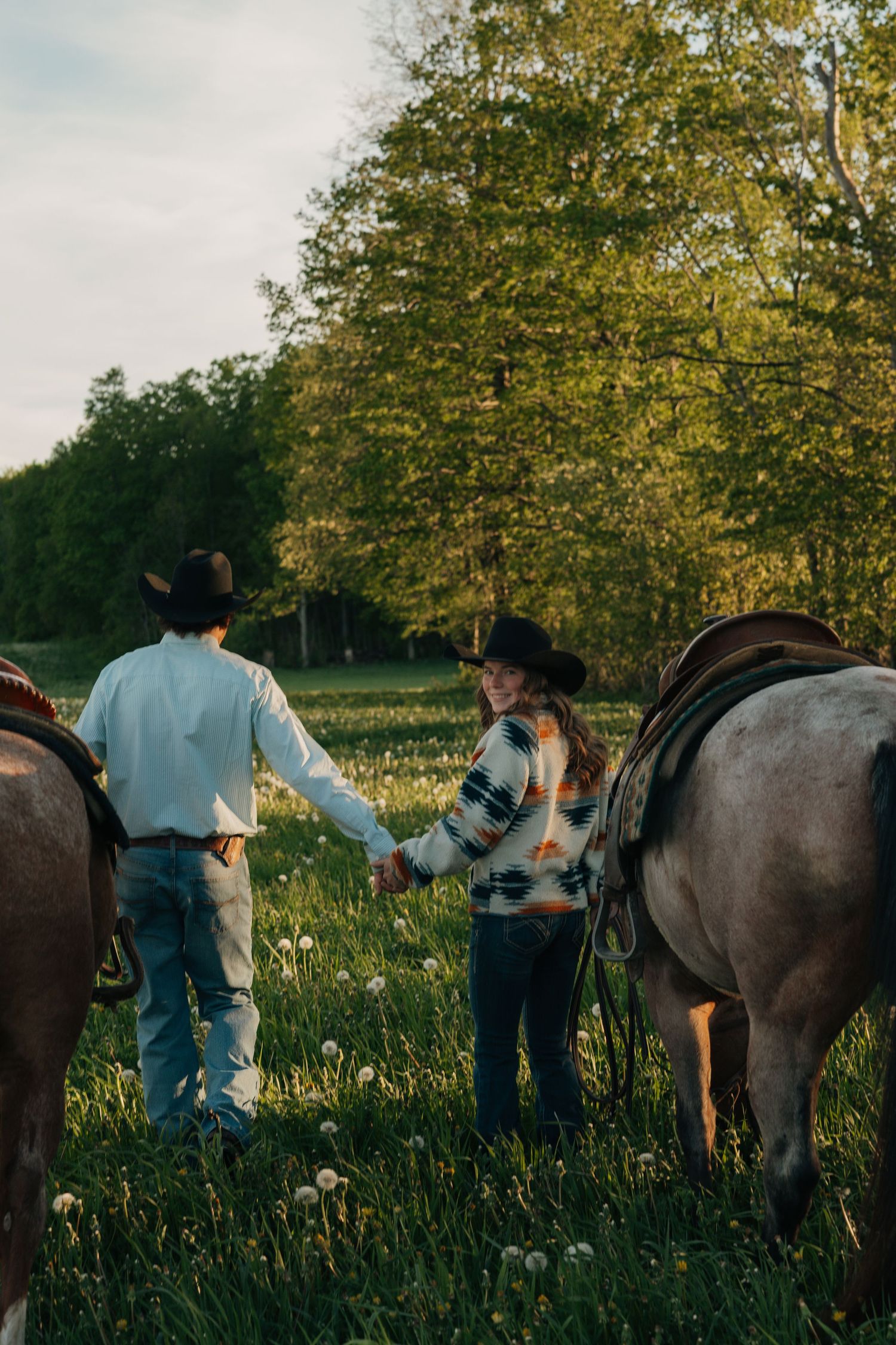 A couple holds hands while walking with horses through a lush green meadow at sunset.