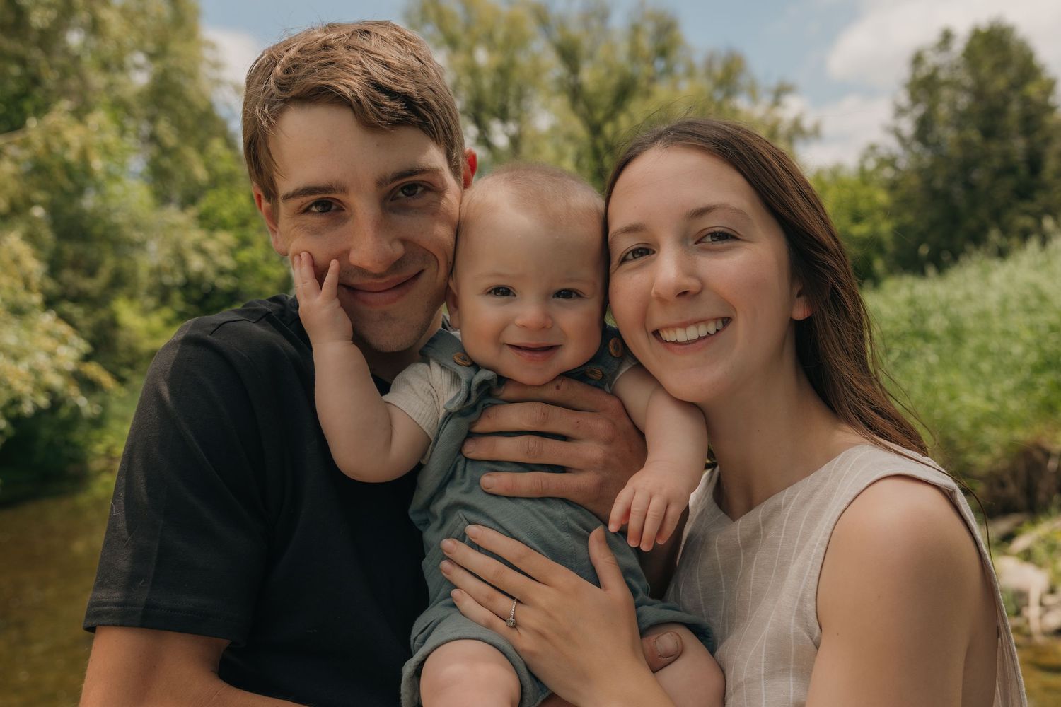 Happy young family with smiling parents holding their laughing baby outdoors near a river on a sunny day.