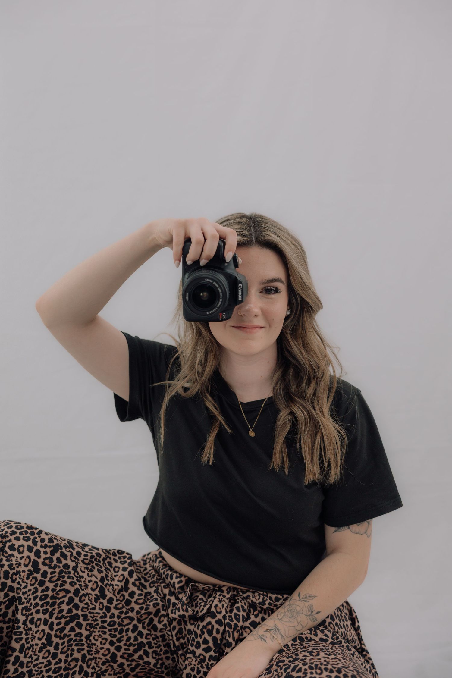 Blonde woman holding a camera up to her face, wearing black top and leopard print skirt.