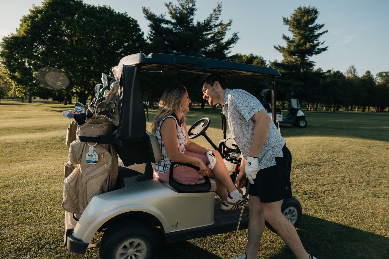 A young couple shares a flirtatious moment beside a golf cart on a sunny golf course.