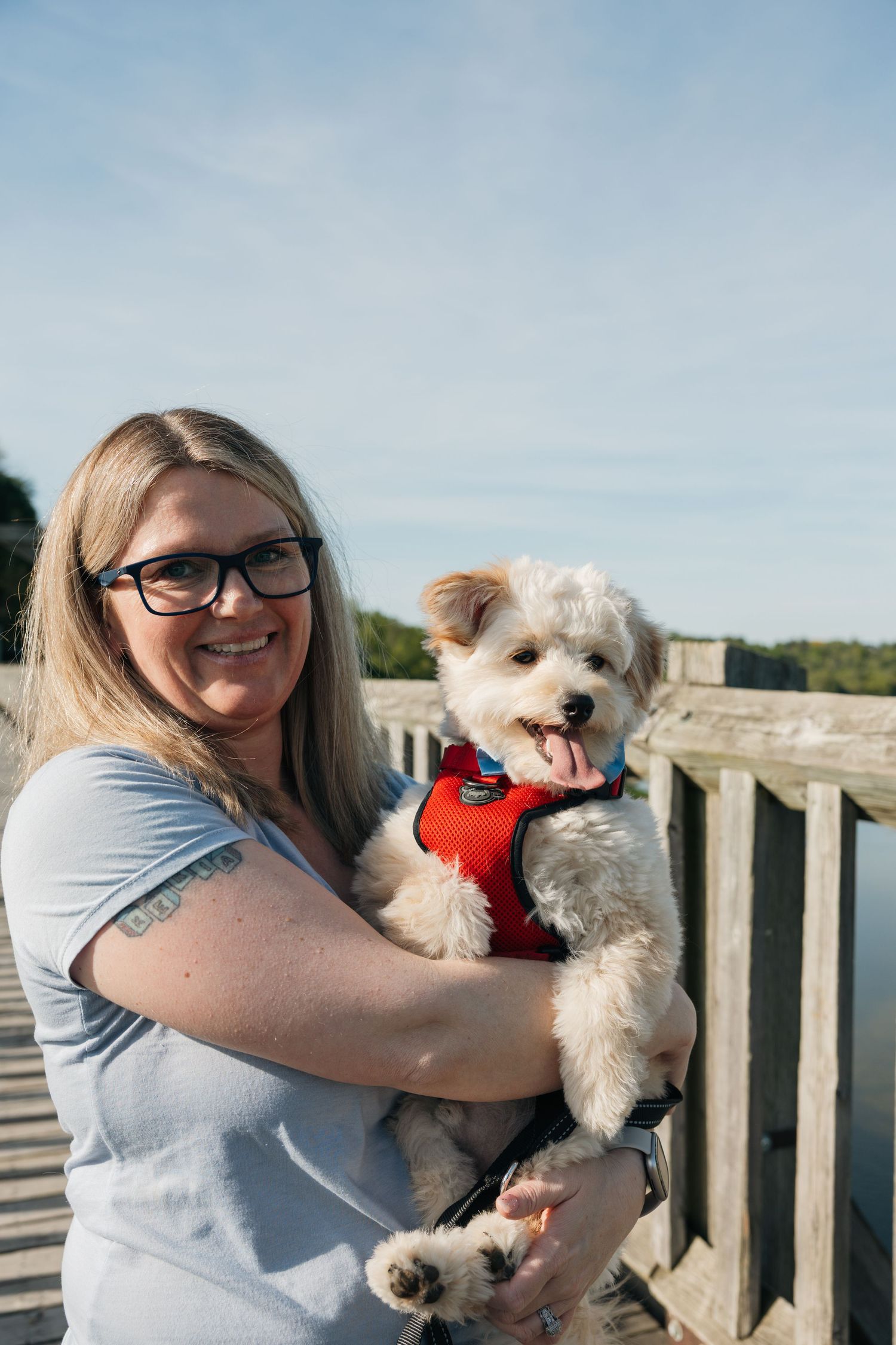 Smiling blonde woman holding fluffy white dog in red harness on wooden bridge.
