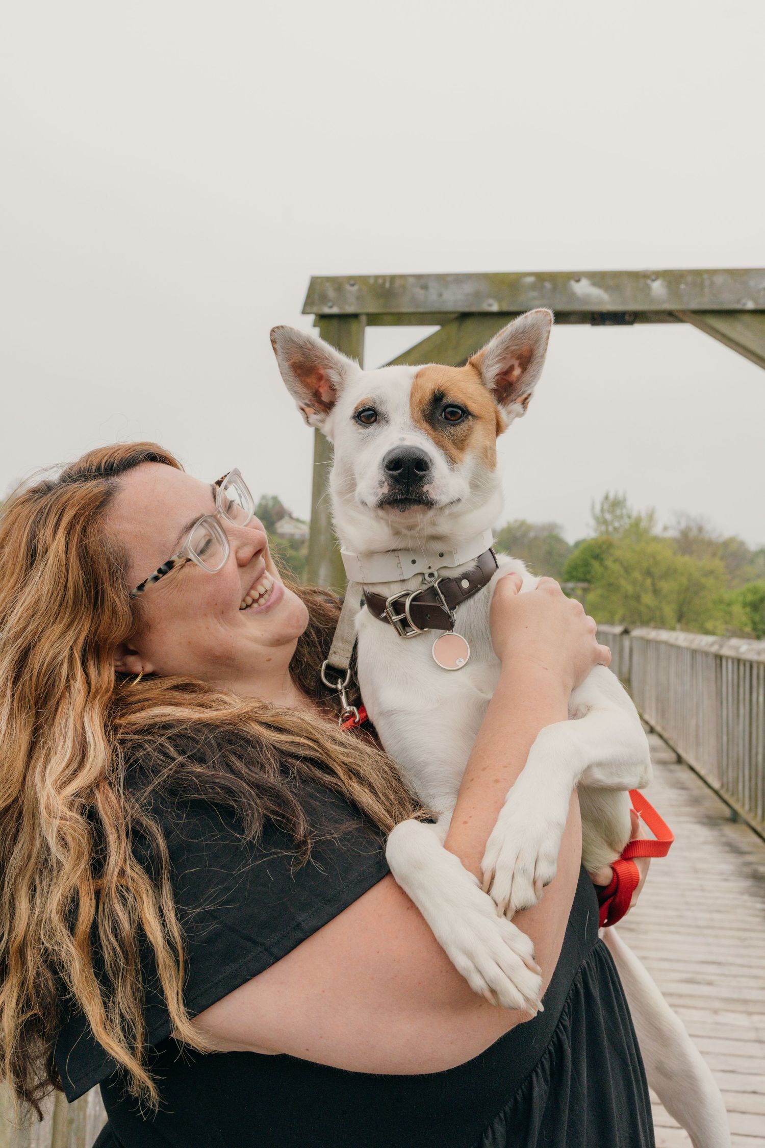 Smiling woman with glasses holds white and tan dog on wooden bridge outdoors.
