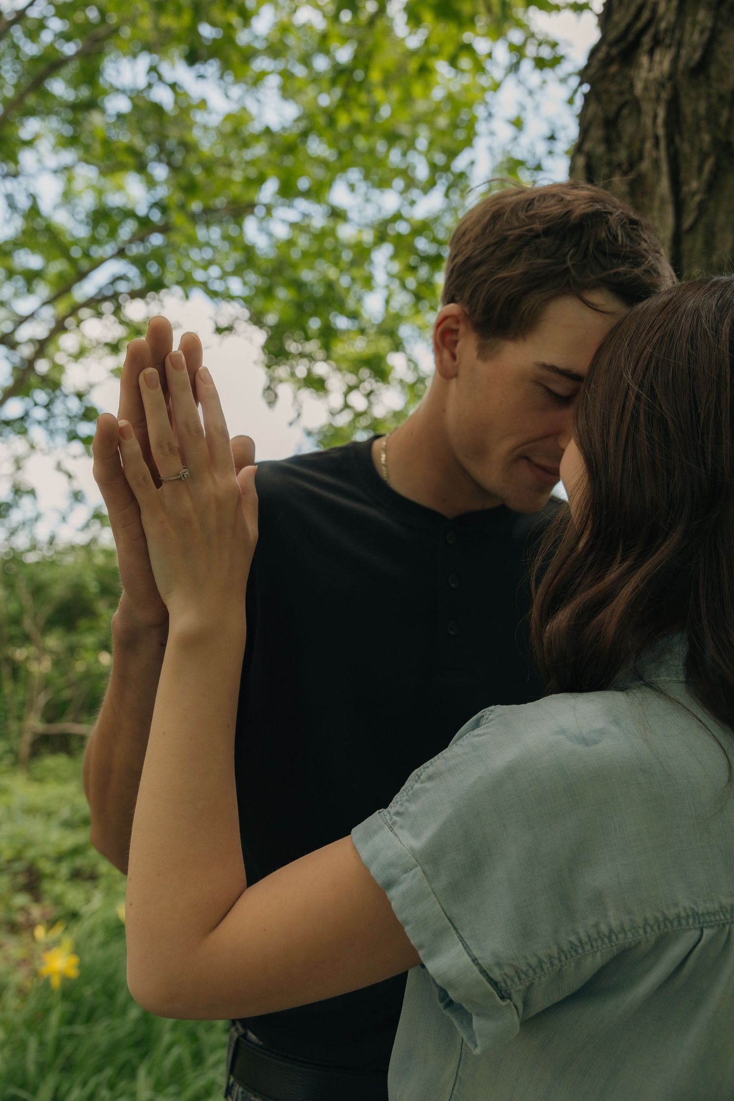 A romantic couple shares an intimate moment pressing hands together near a tree in a lush green outdoor setting.