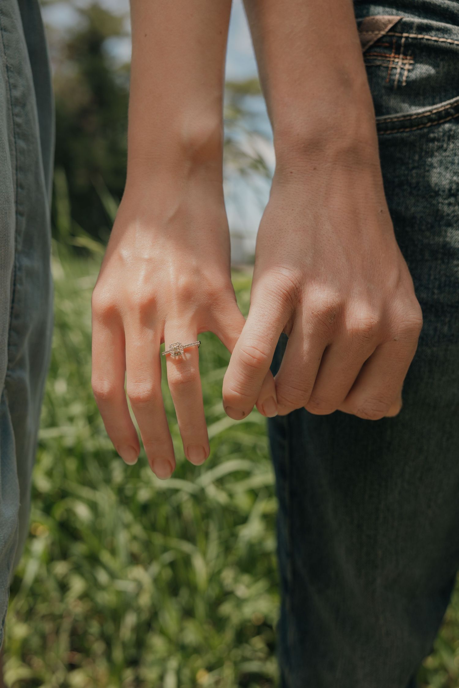 Close-up of two people gently linking pinky fingers, one wearing an engagement ring, in a grassy field.