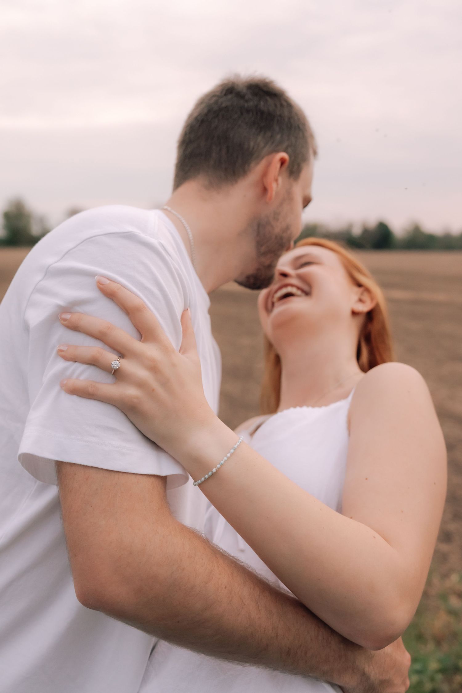 Joyful couple embracing and laughing together in an open field, woman wearing engagement ring and white dress.