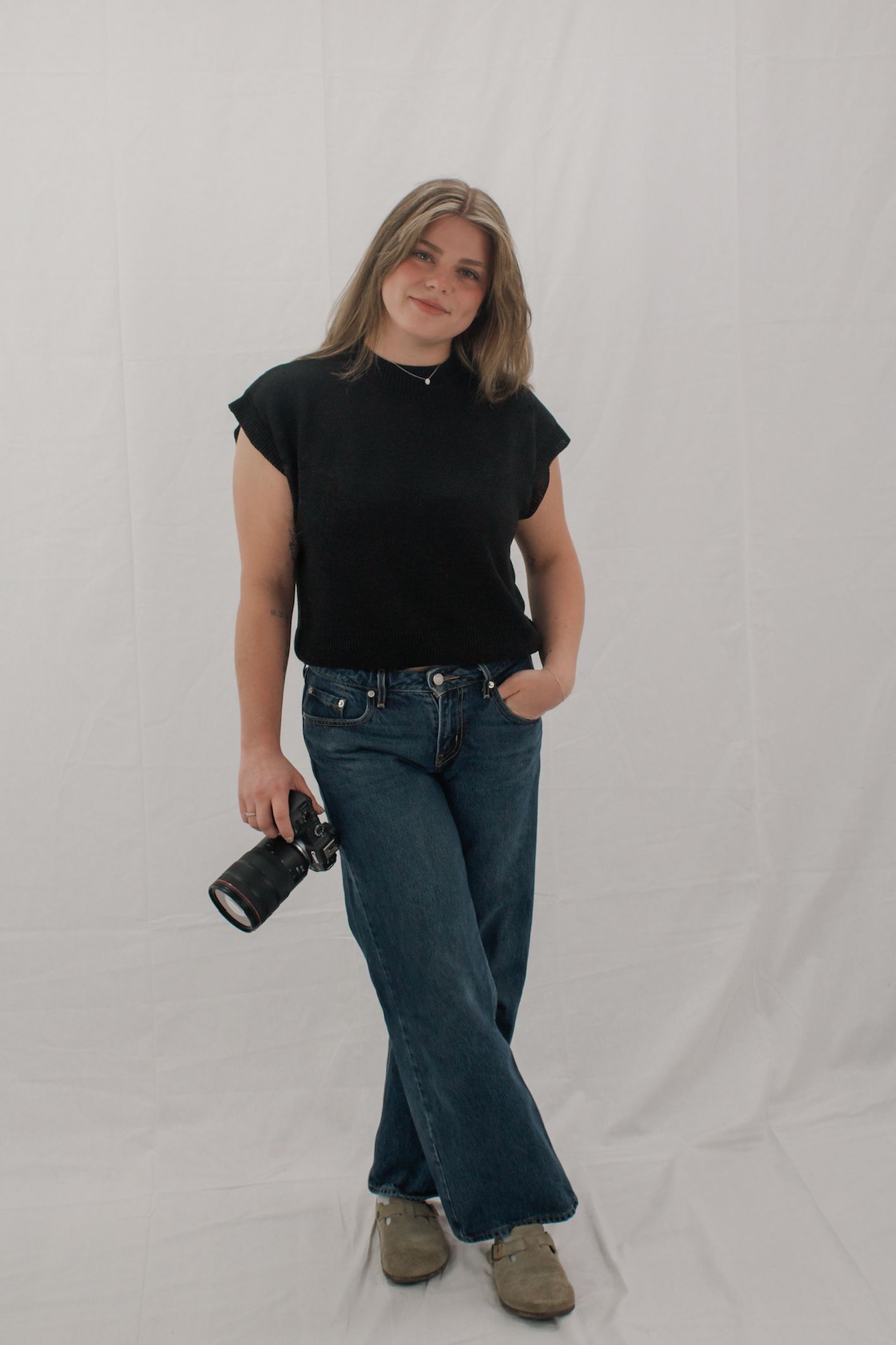 Young blonde female photographer standing confidently holding a camera camera in black top and wide-leg jeans.