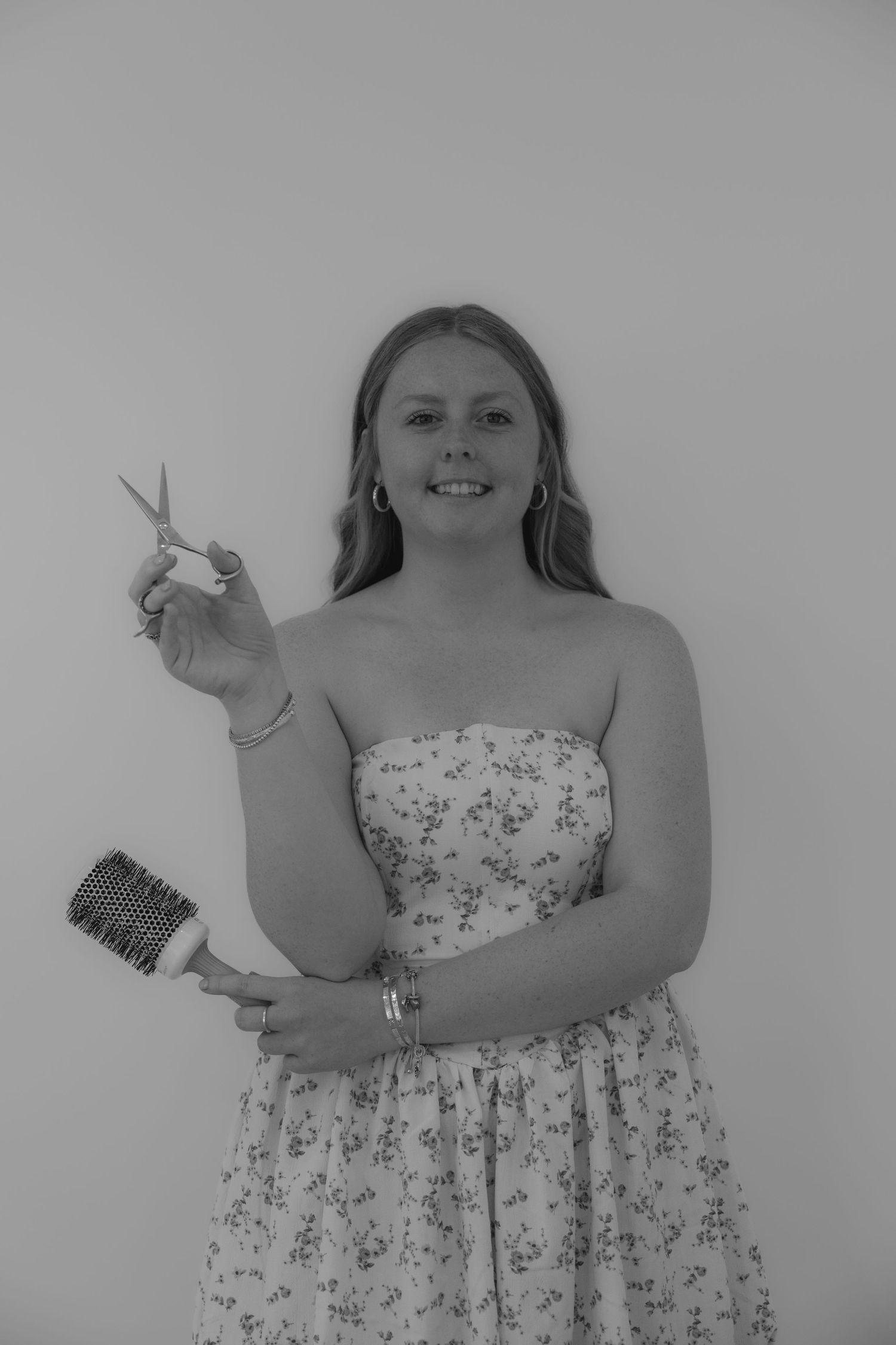 Black and white portrait of young woman in strapless floral dress holding scissors and round hair brush.