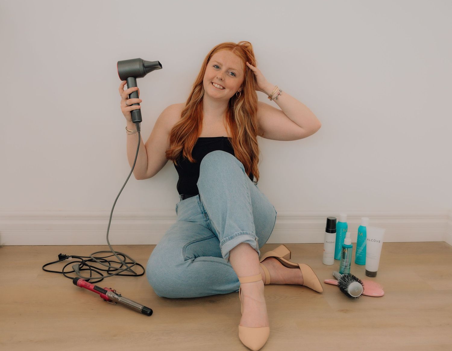 Redhead woman sitting on floor holding hair dryer surrounded by styling tools and hair products.