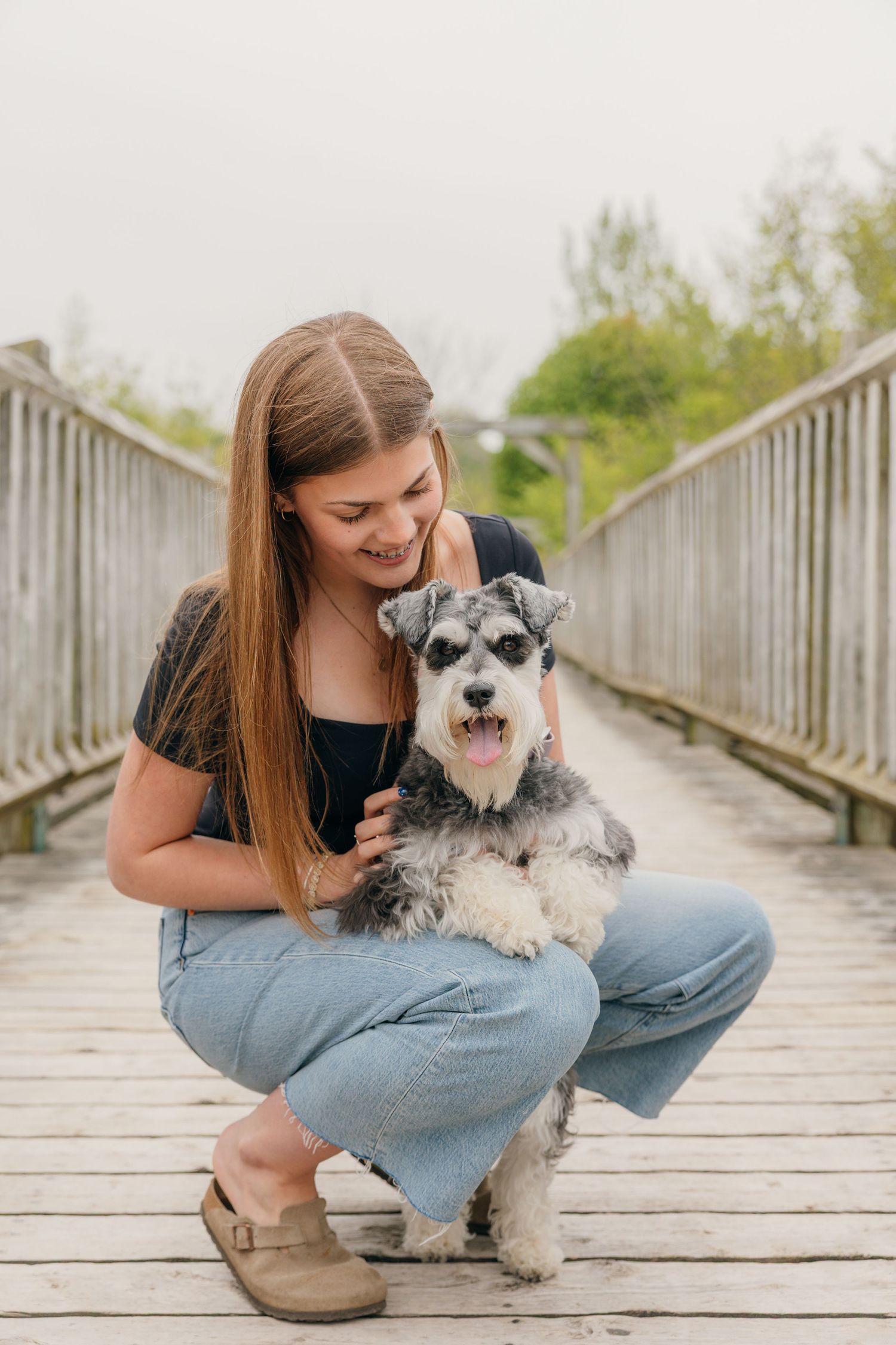 Smiling teenage girl crouching on wooden bridge holding a miniature schnauzer dog.