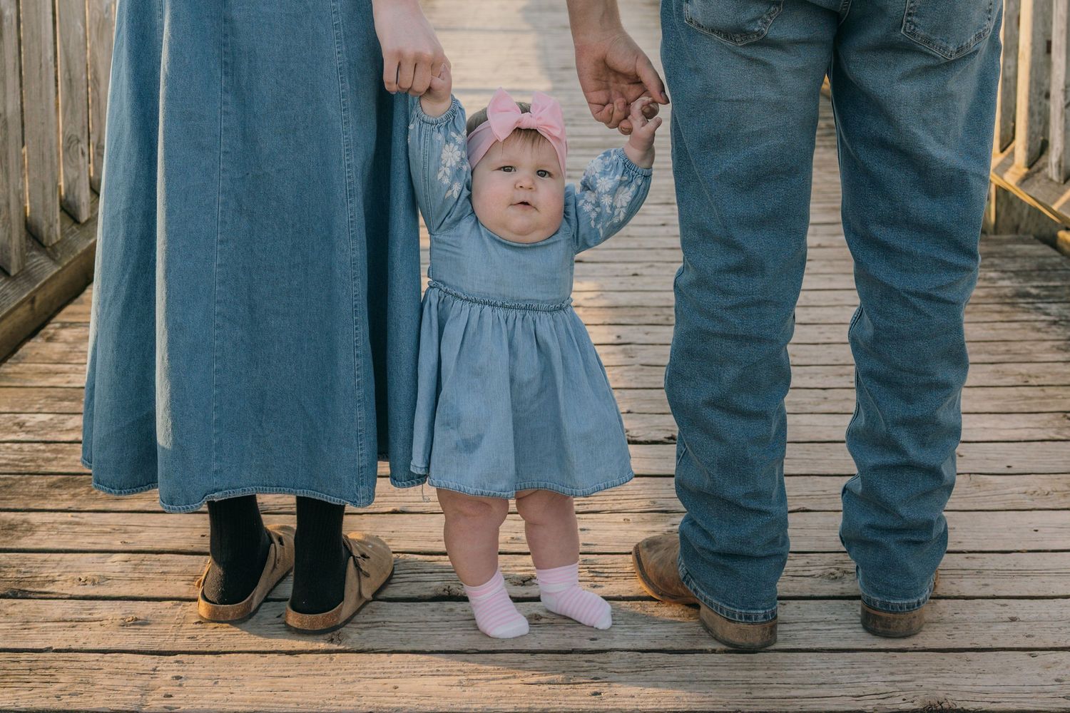 Baby girl in blue dress standing between parents on a wooden bridge.