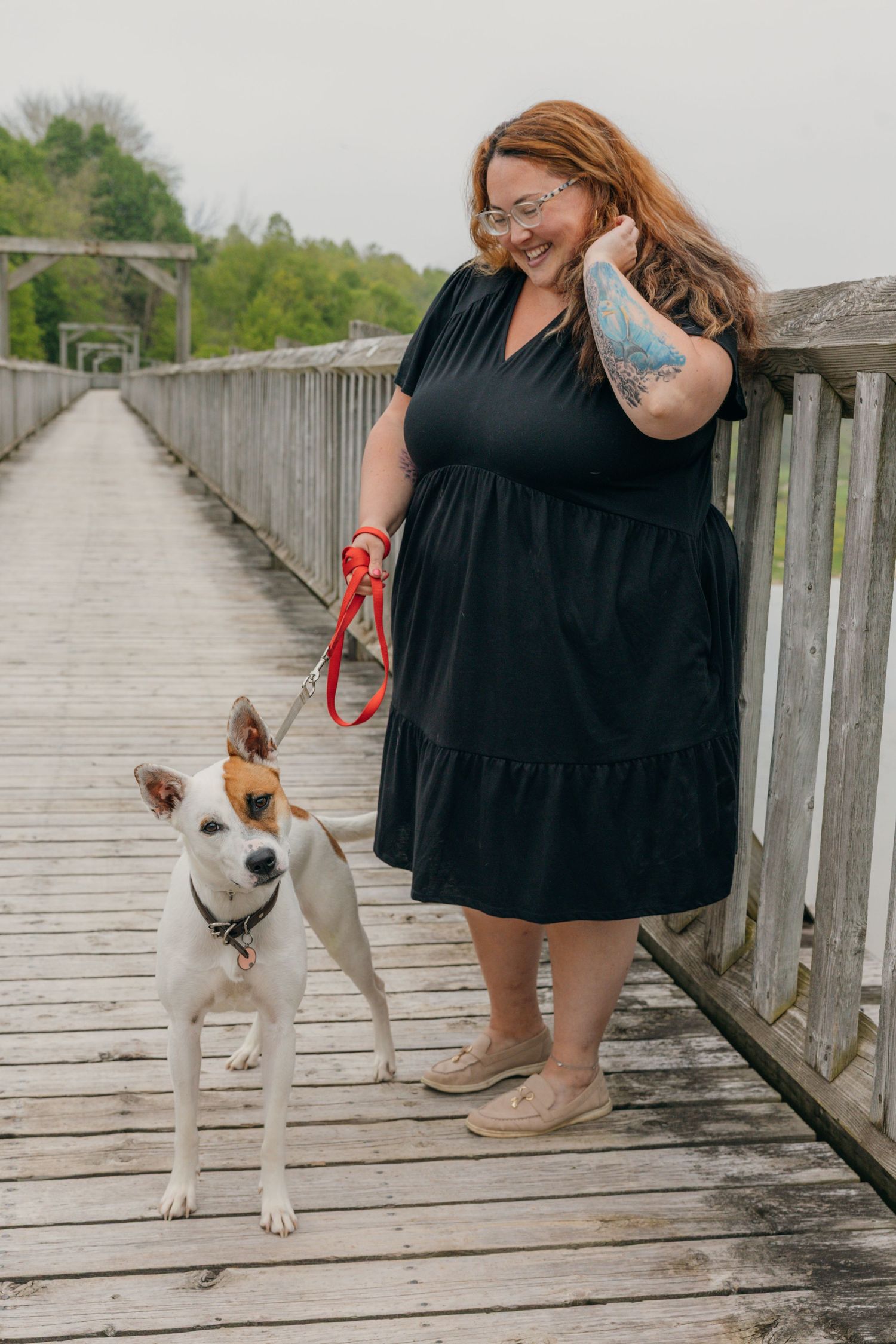 Woman in black dress walks dog on red leash on wooden bridge.