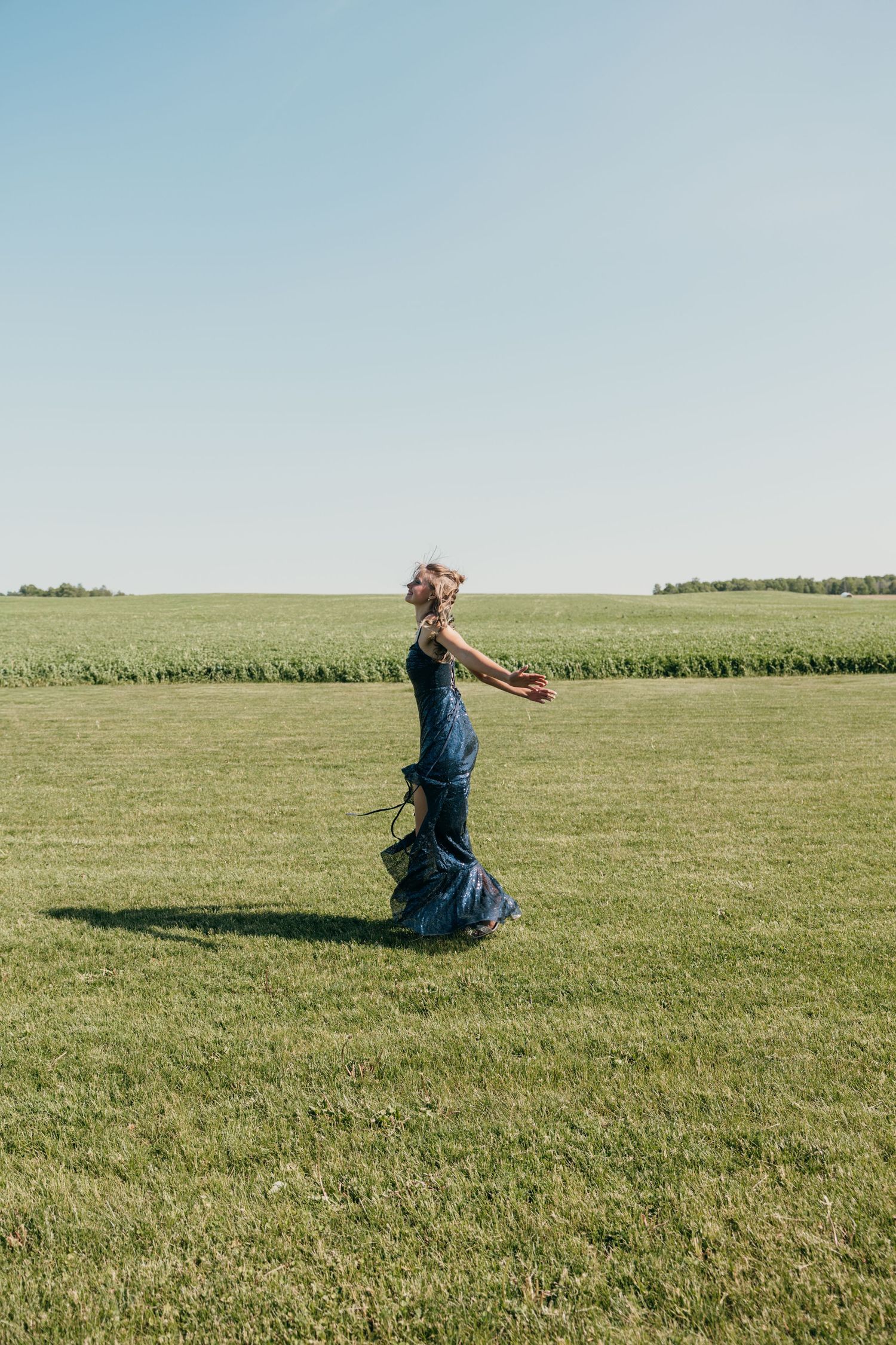 Woman in flowing blue sequin gown walking through vast green field under clear blue sky.
