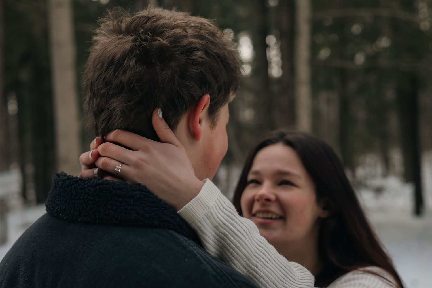A happy woman with an engagement ring embraces a man in a snowy forest setting.