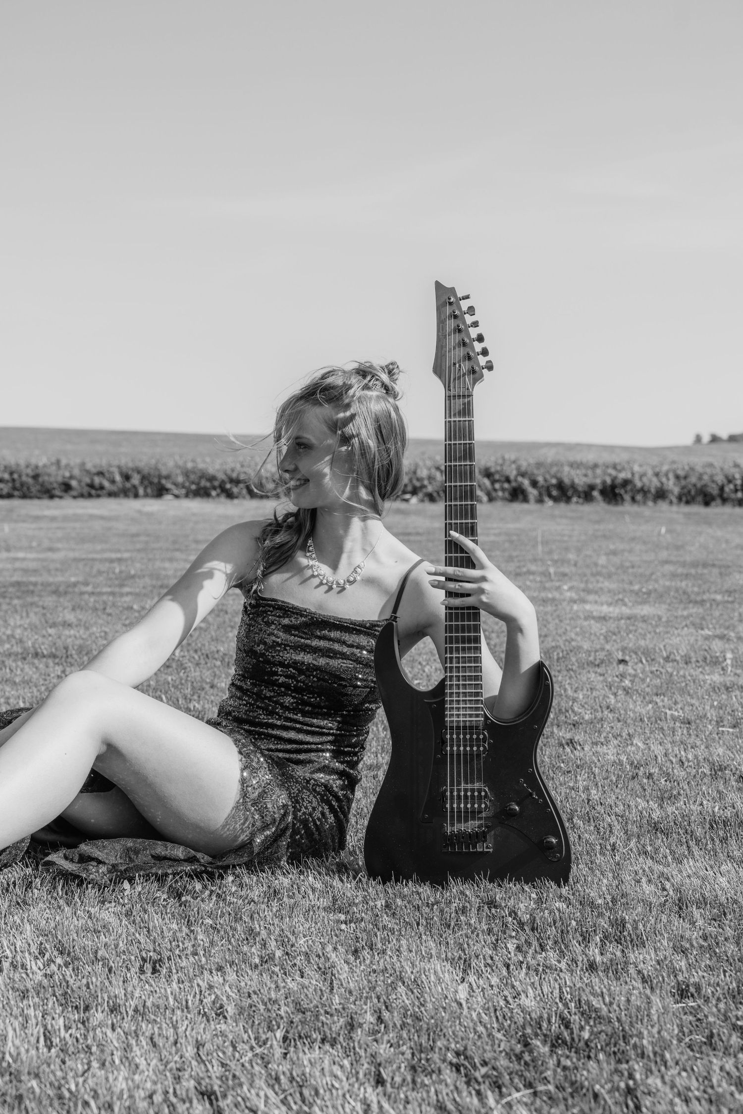 Black and white photo of smiling young woman in sequin dress sitting on grass with electric guitar.