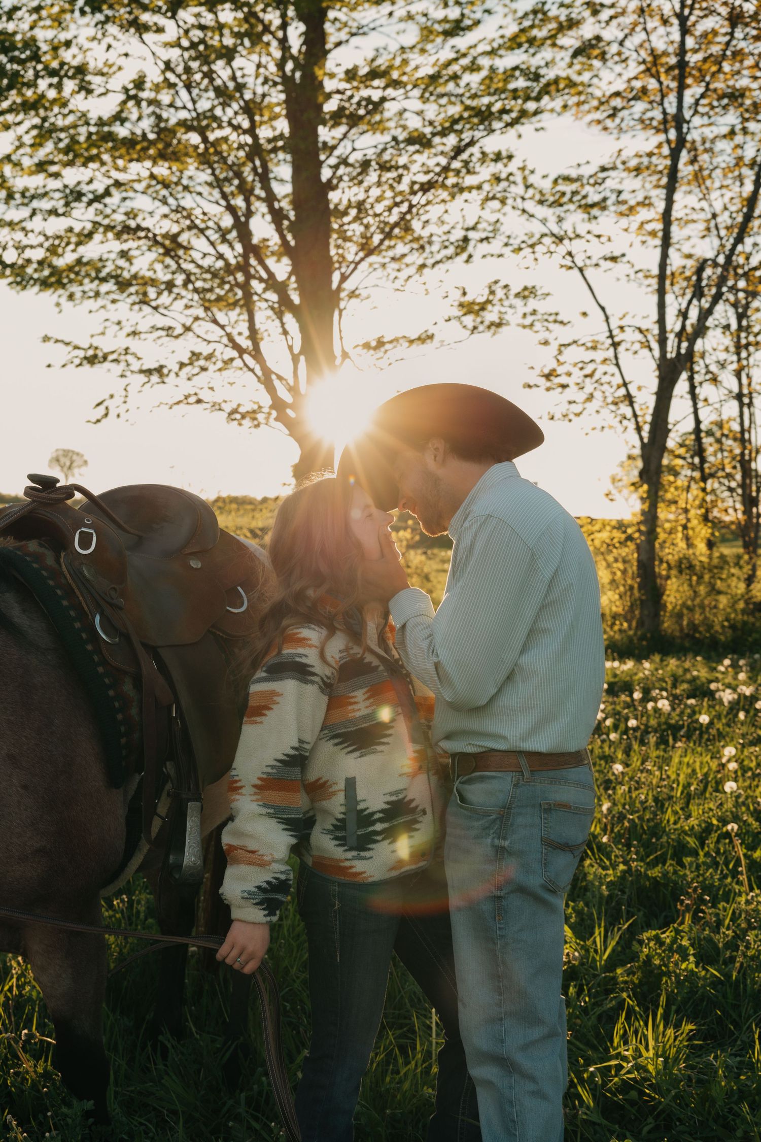 A couple shares a tender moment beside a horse with golden sunset light streaming through trees.