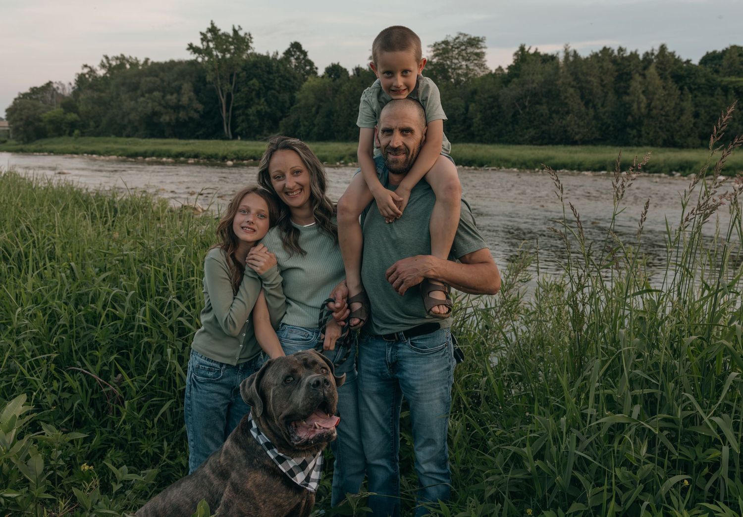 Smiling family of four with a gray dog posing by a river surrounded by green vegetation.