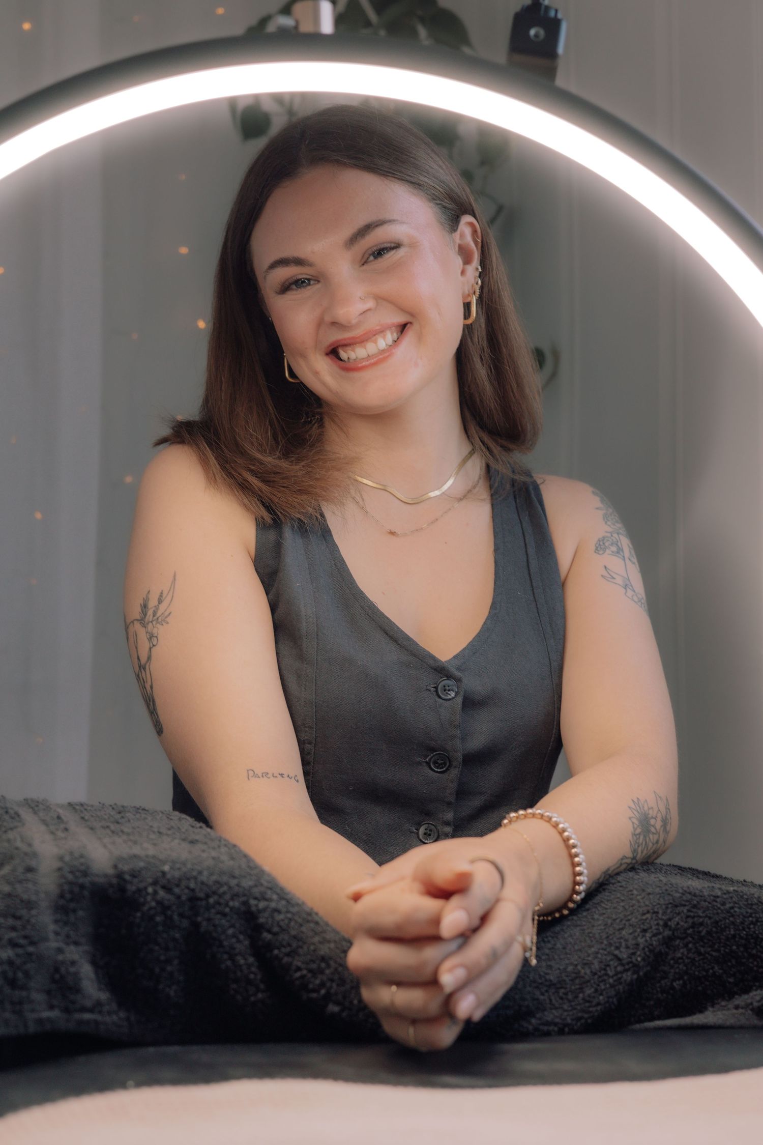 Smiling young woman with tattoos sits under ring light in gray vest top.