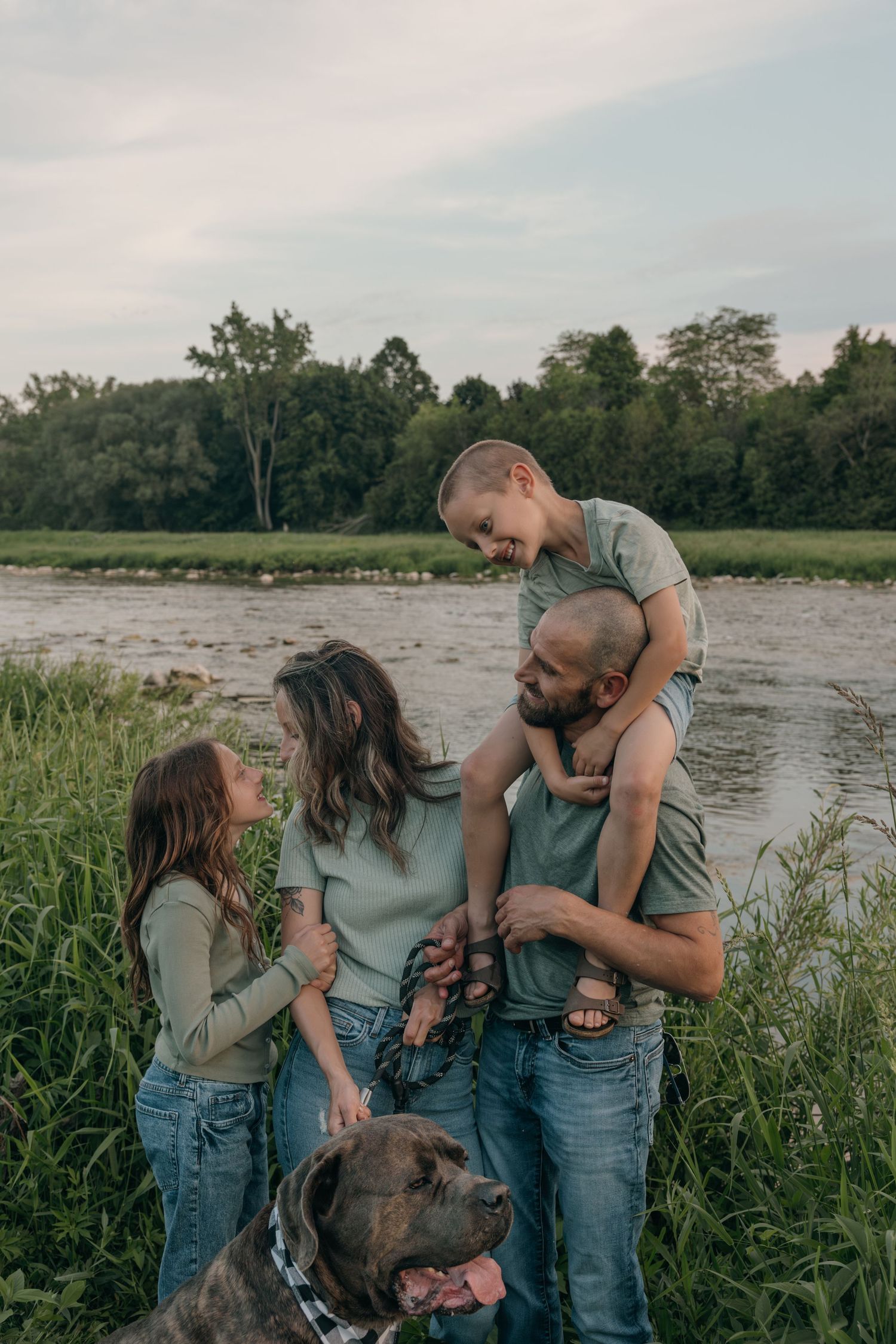Joyful family of four with a brindle dog laughing together by a scenic river in tall grass.