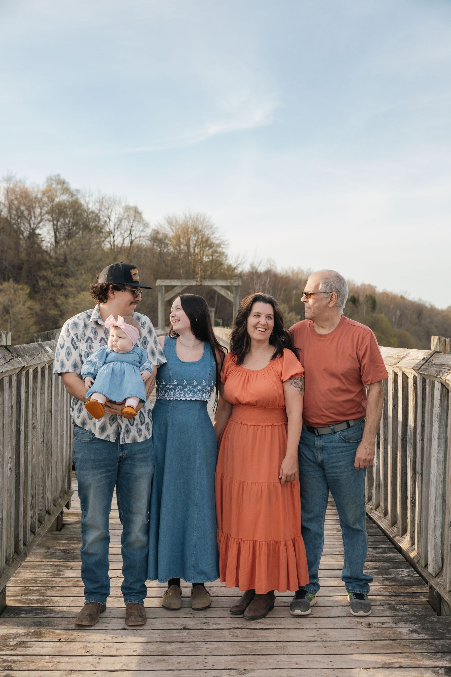 Family of four with a baby poses on a wooden bridge outdoors on a sunny day.