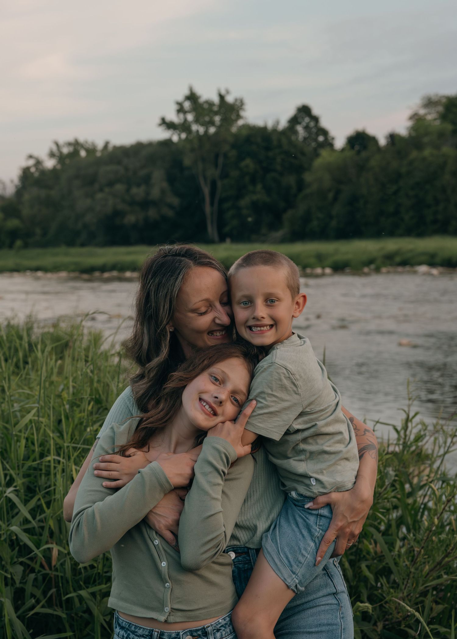 Mother hugging her two smiling children by a river surrounded by lush green trees and grass.