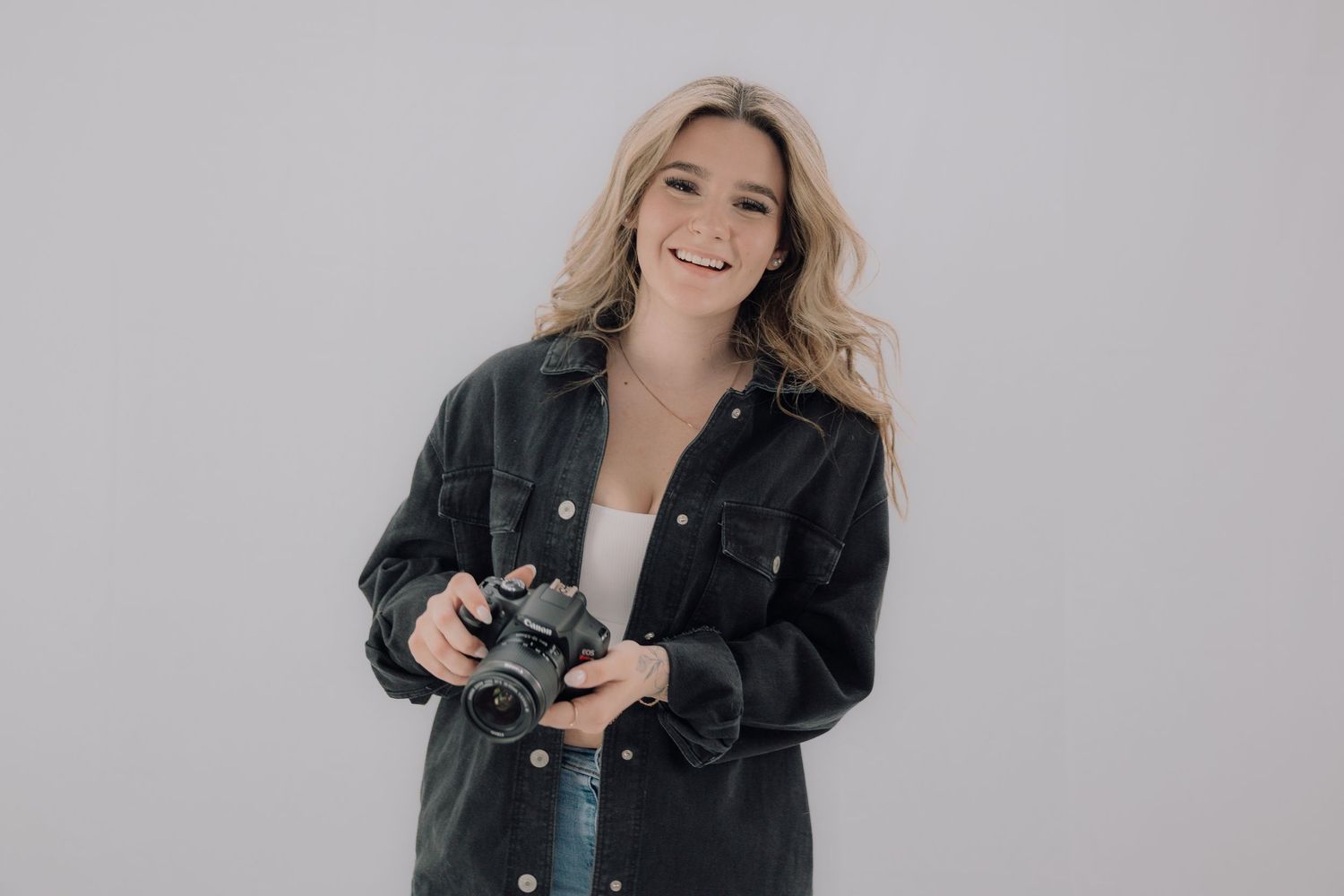 Smiling blonde woman holding a camera wearing a black denim jacket against a gray background.