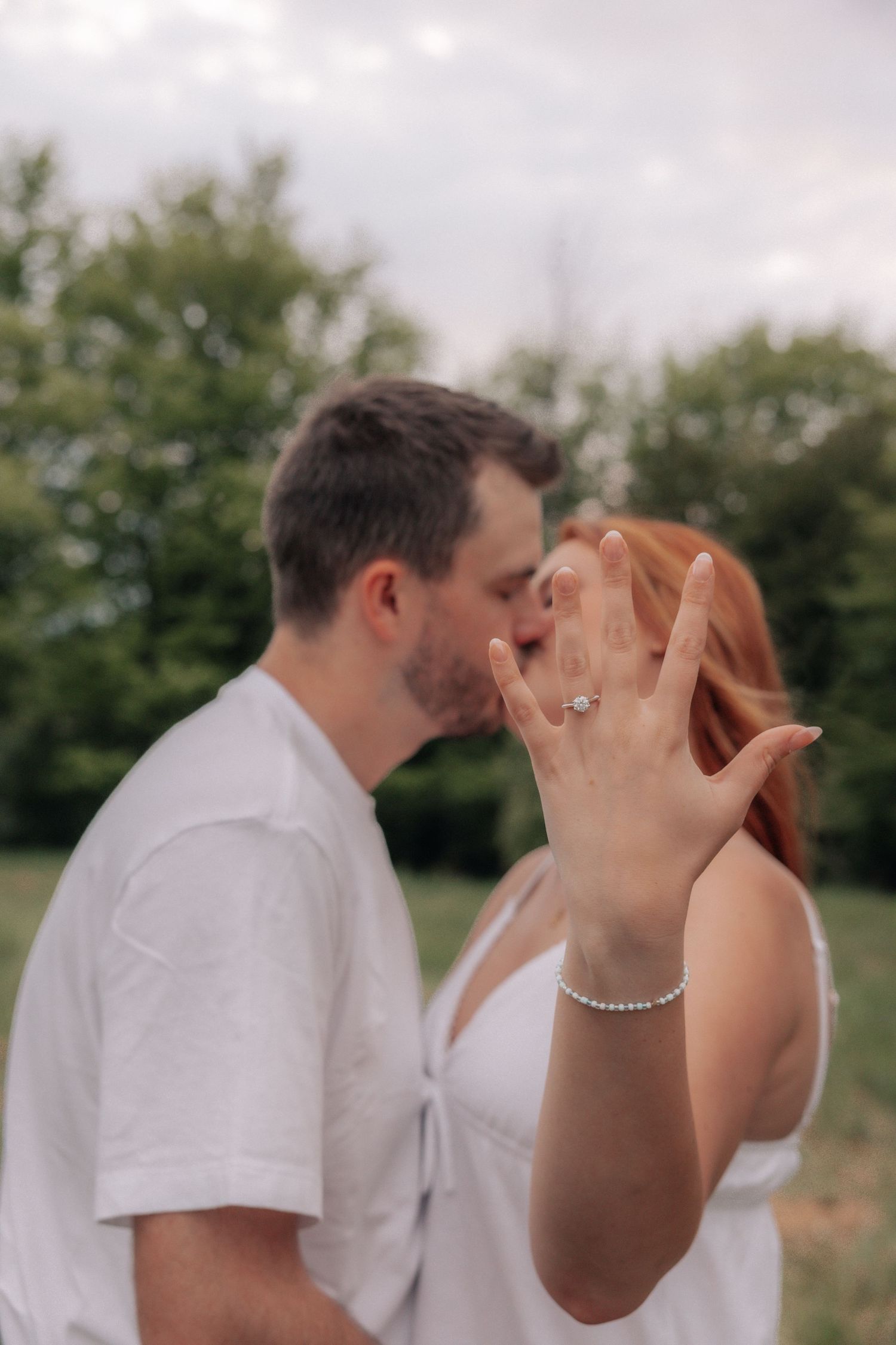 Engaged couple kissing outdoors while woman displays her diamond engagement ring to camera.