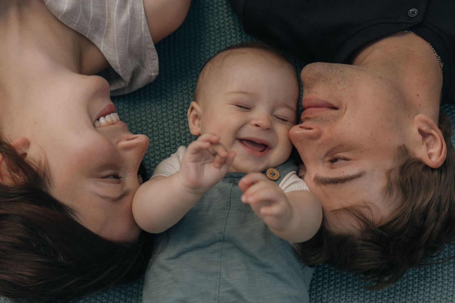 Two parents lying down with their laughing baby between them, sharing a joyful family moment.