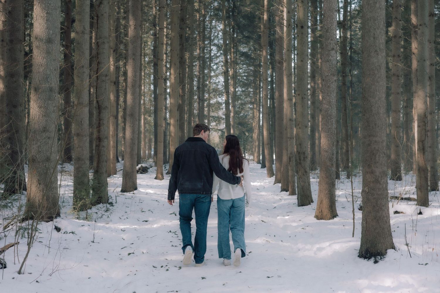 Couple walking hand in hand through a snowy winter forest with tall trees.