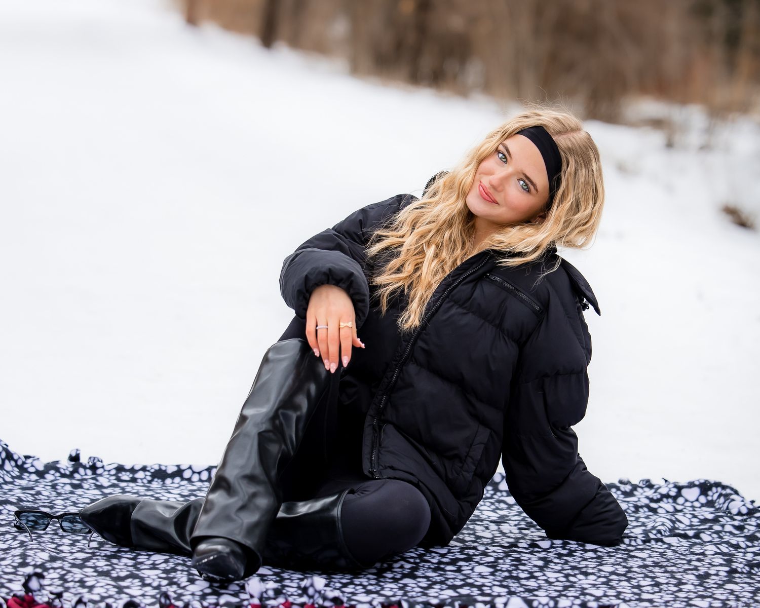 Blonde woman in black puffer jacket and boots sitting on blanket in snowy winter landscape.
