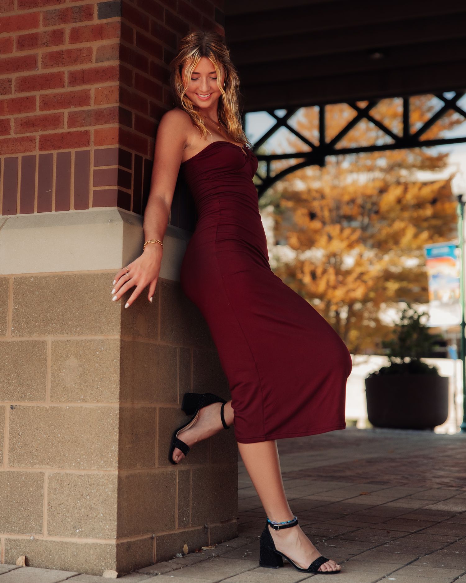 Person in burgundy dress poses playfully against brick building exterior.