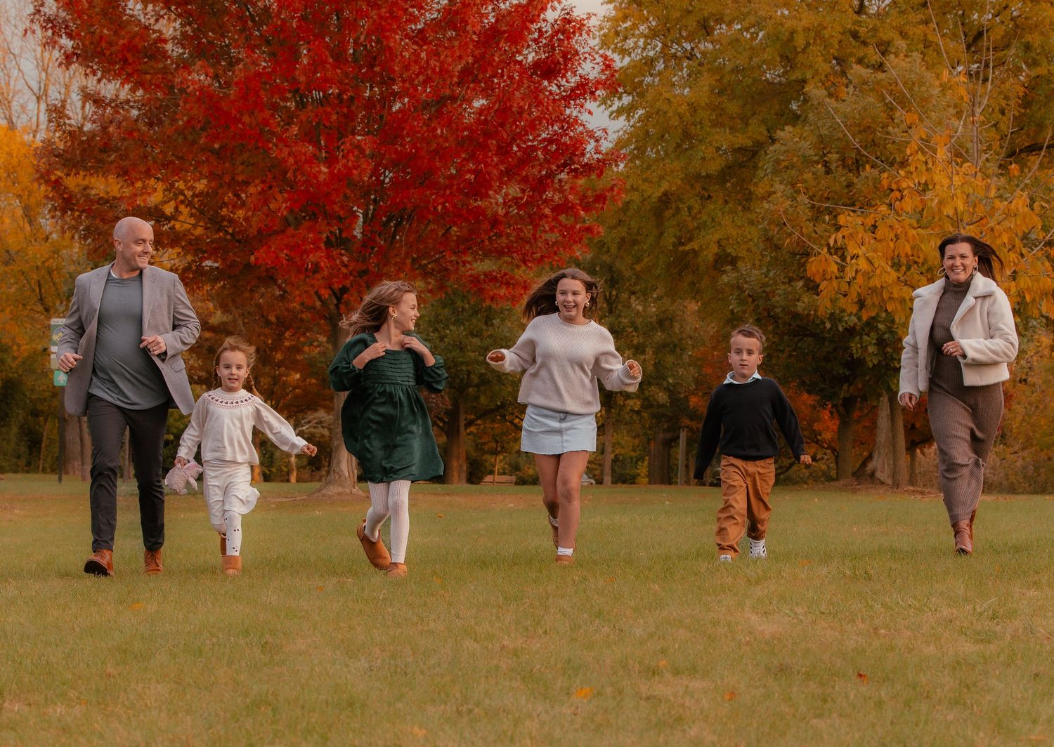 Family members run across field with vibrant red and orange fall foliage.