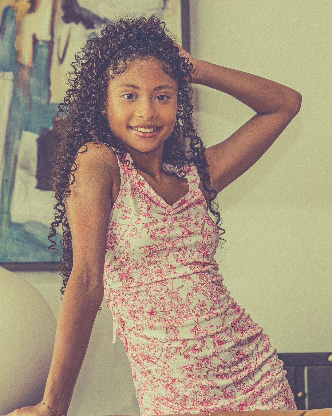 Young girl with curly hair smiling in a pink floral dress, posing confidently indoors near colorful wall art.