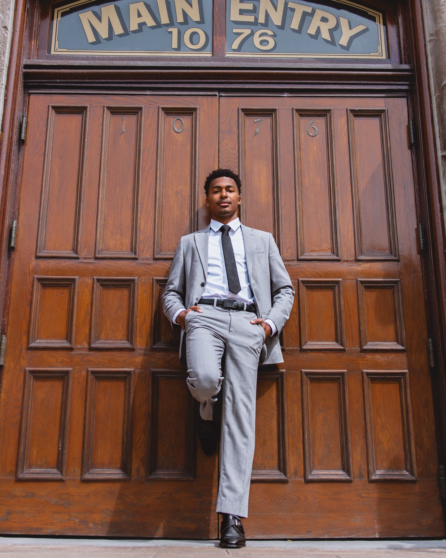Young man in gray suit poses confidently against ornate wooden Main Entry doors, number 1076.