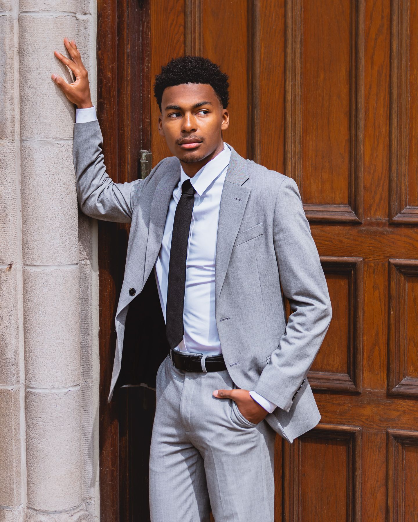 Young man in gray suit and black tie leaning against ornate wooden door in professional portrait photo.
