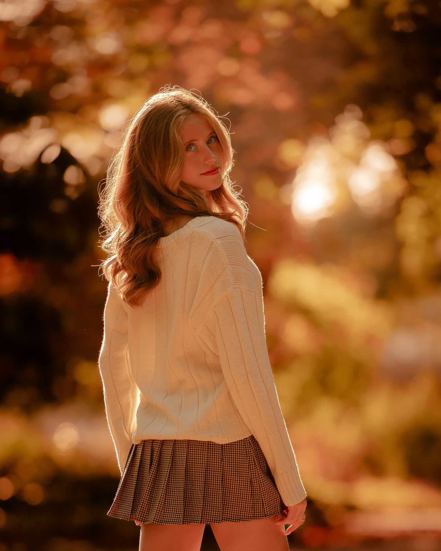 Young woman in white sweater and plaid skirt poses outdoors among autumn foliage in warm golden light.